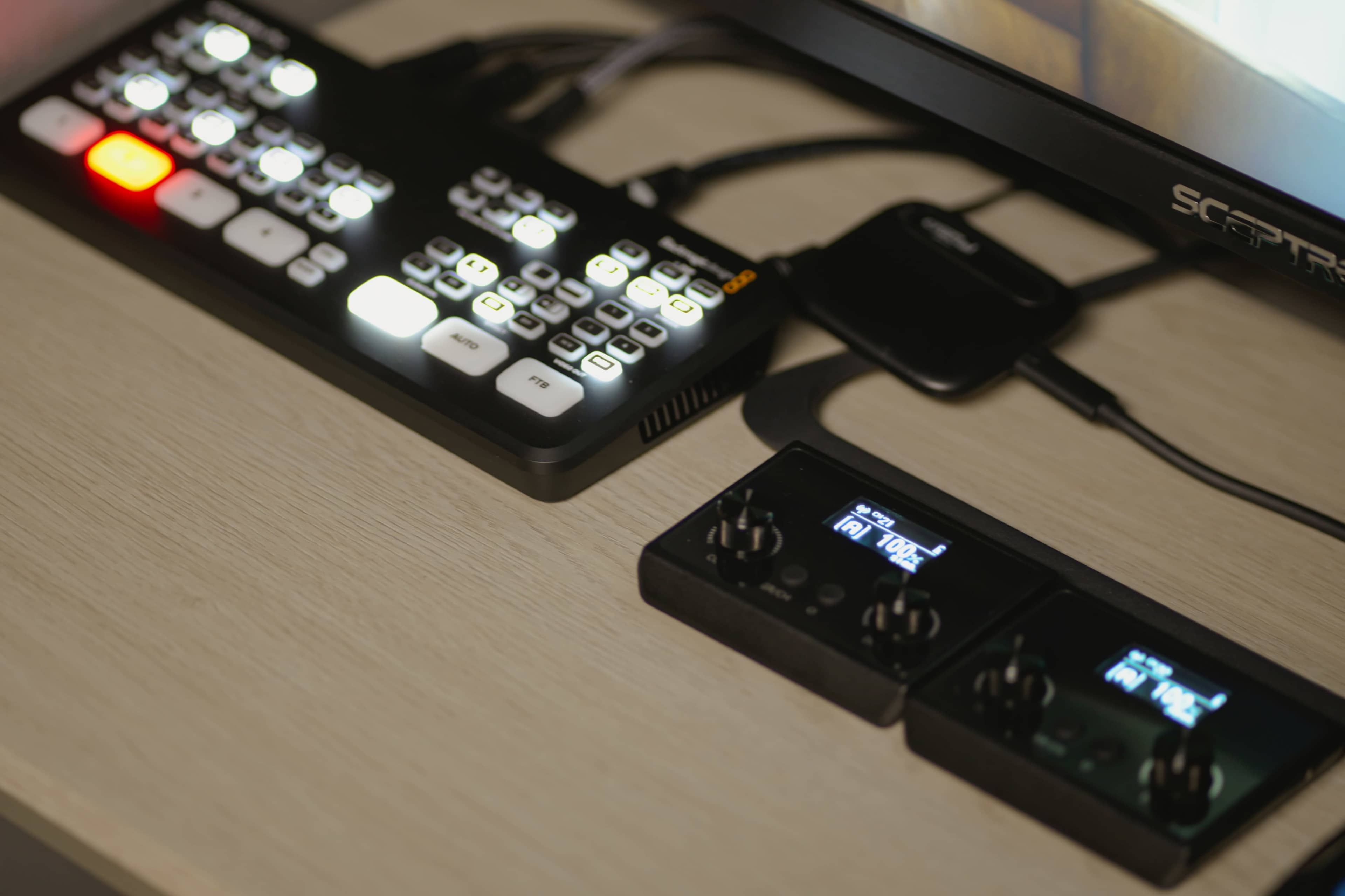 The image shows a digital control panel with backlit buttons and two audio interface controllers on a desk next to a computer monitor.