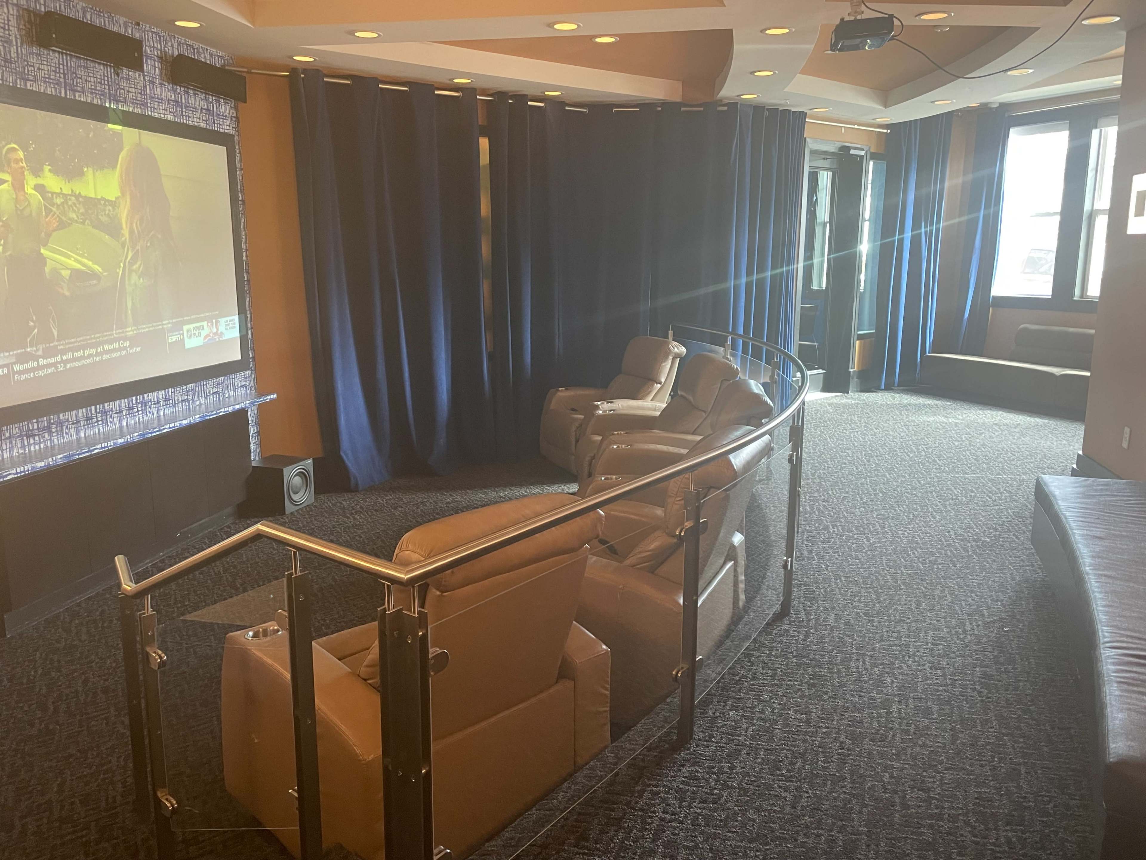 A home theater setup featuring plush reclining chairs, a large screen displaying a movie, and dimmed lighting with blue curtains.