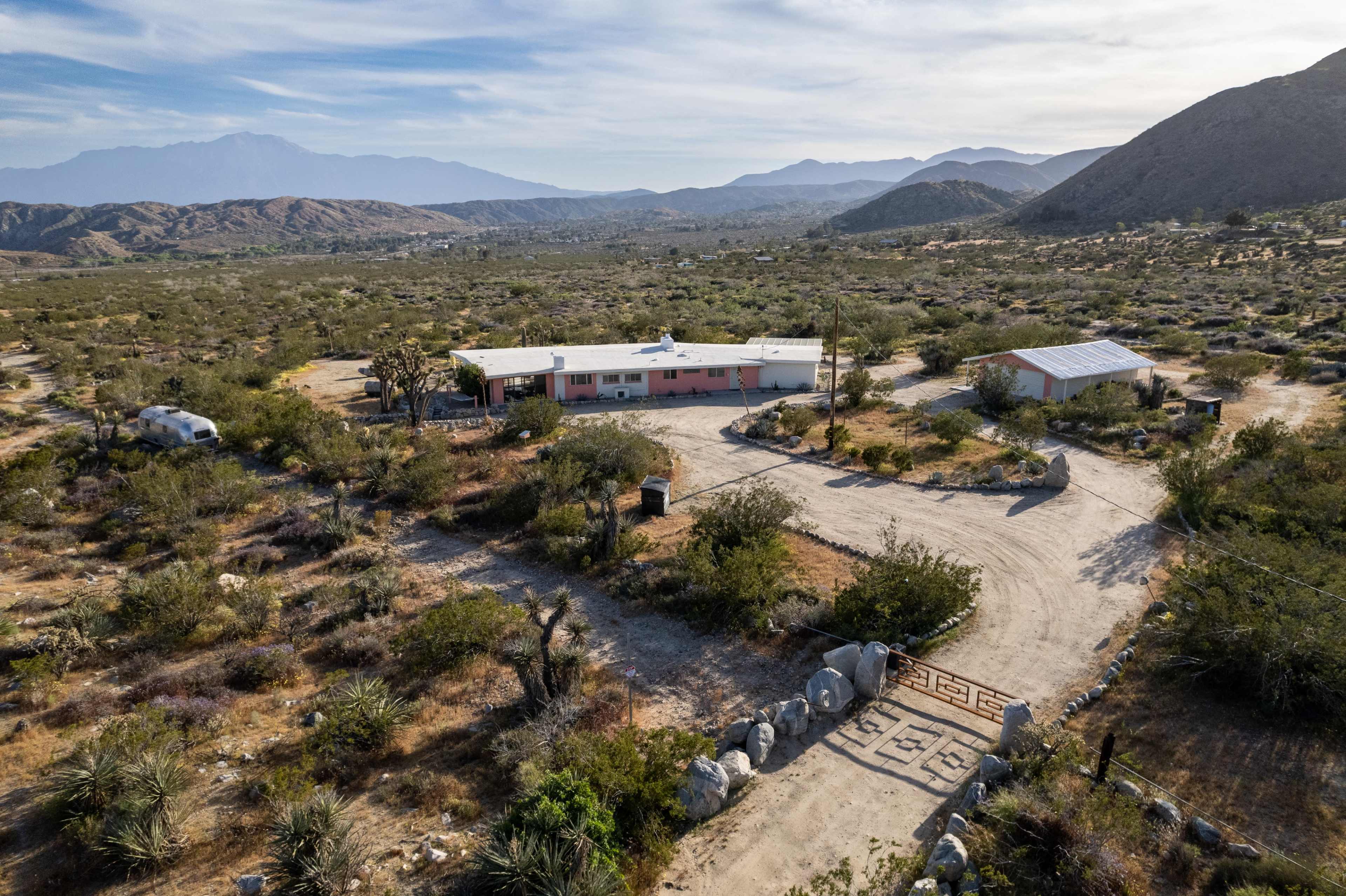 The image shows an aerial view of a ranch-style house situated in a desert landscape, surrounded by hills and sparse vegetation.