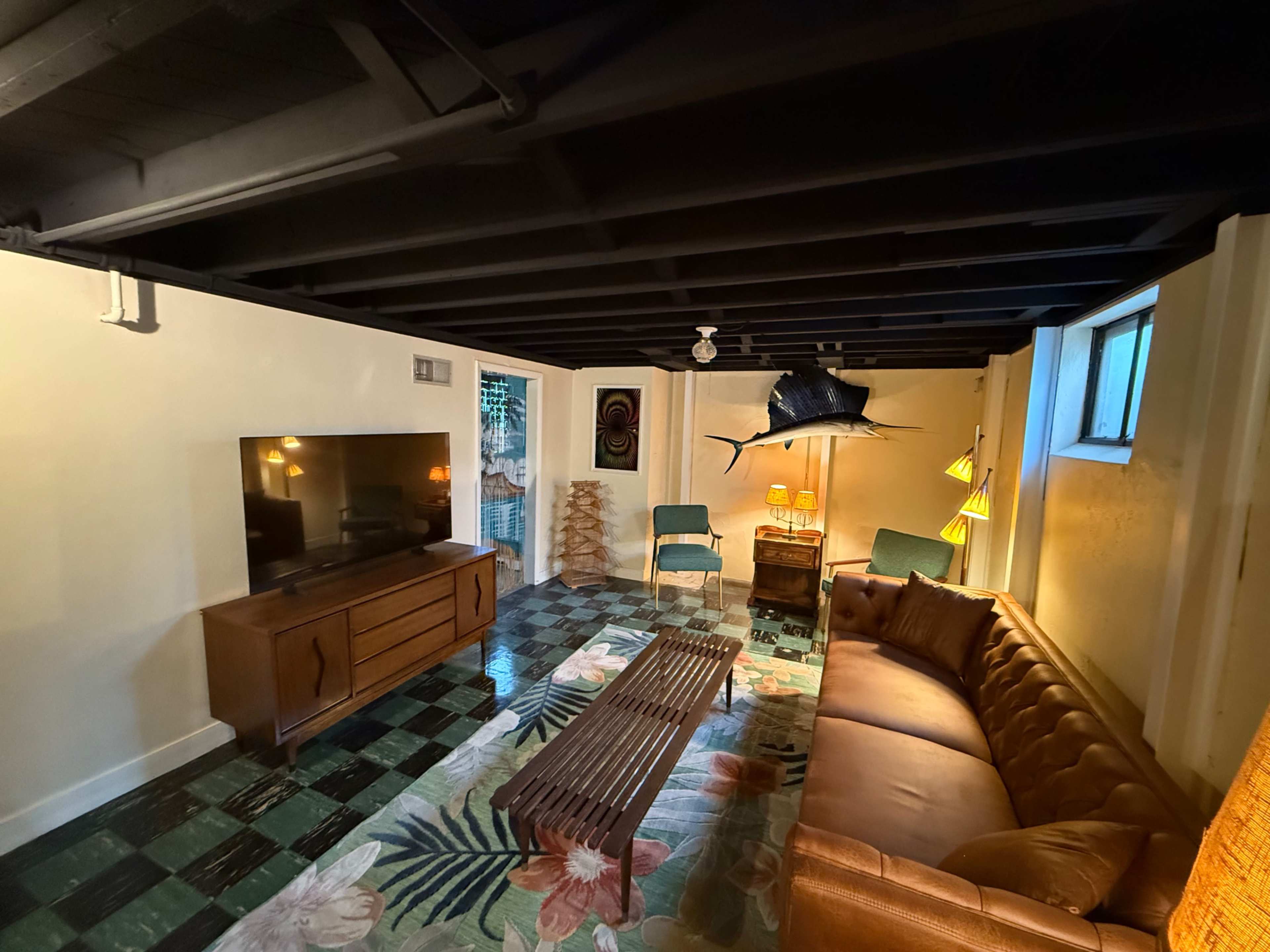 The image shows a cozy basement living area with a brown leather sofa, a wooden coffee table, a large television, a mounted fish on the wall, and two green chairs beside a window.