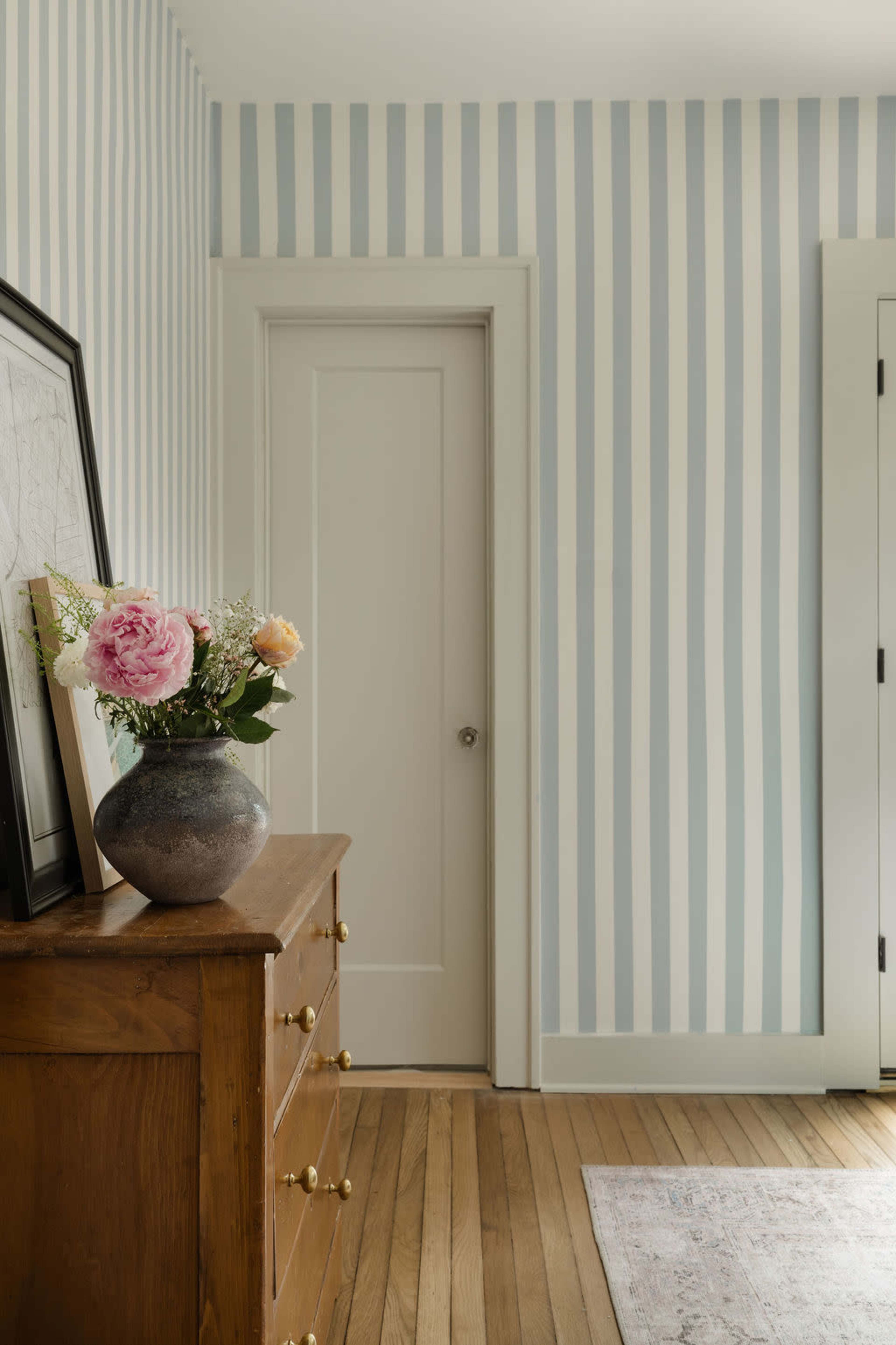The image shows a room with light blue and white striped walls, a wooden dresser with brass knobs, a closed white door, and a vase with flowers on top of the dresser.