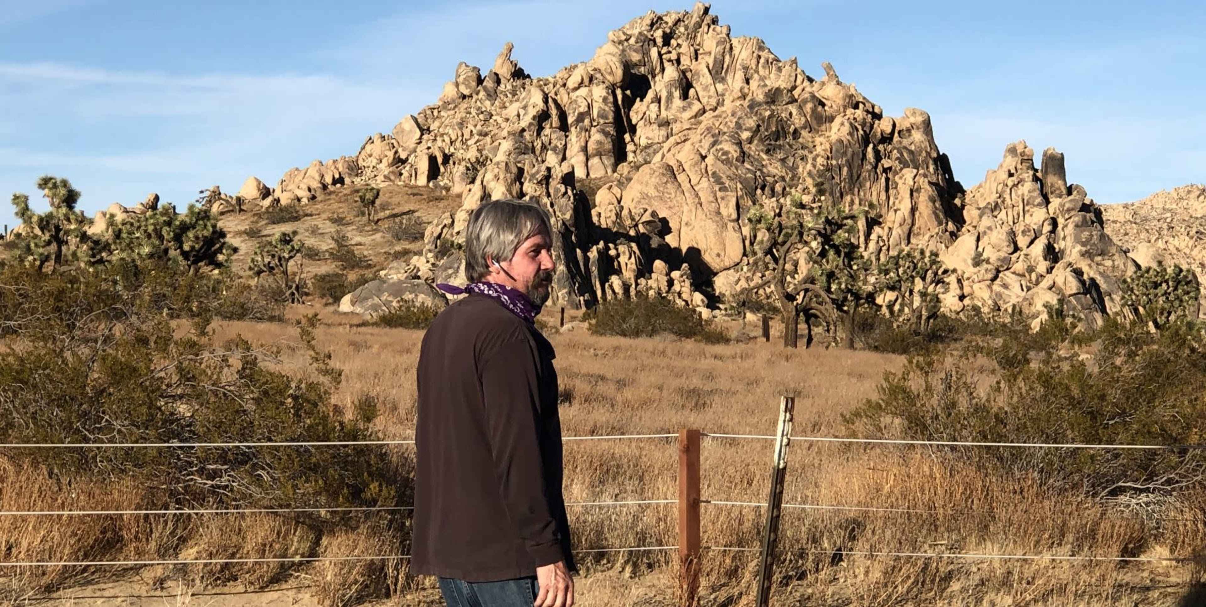 A man stands next to a wooden fence in a desert landscape with large rock formations in the background.
