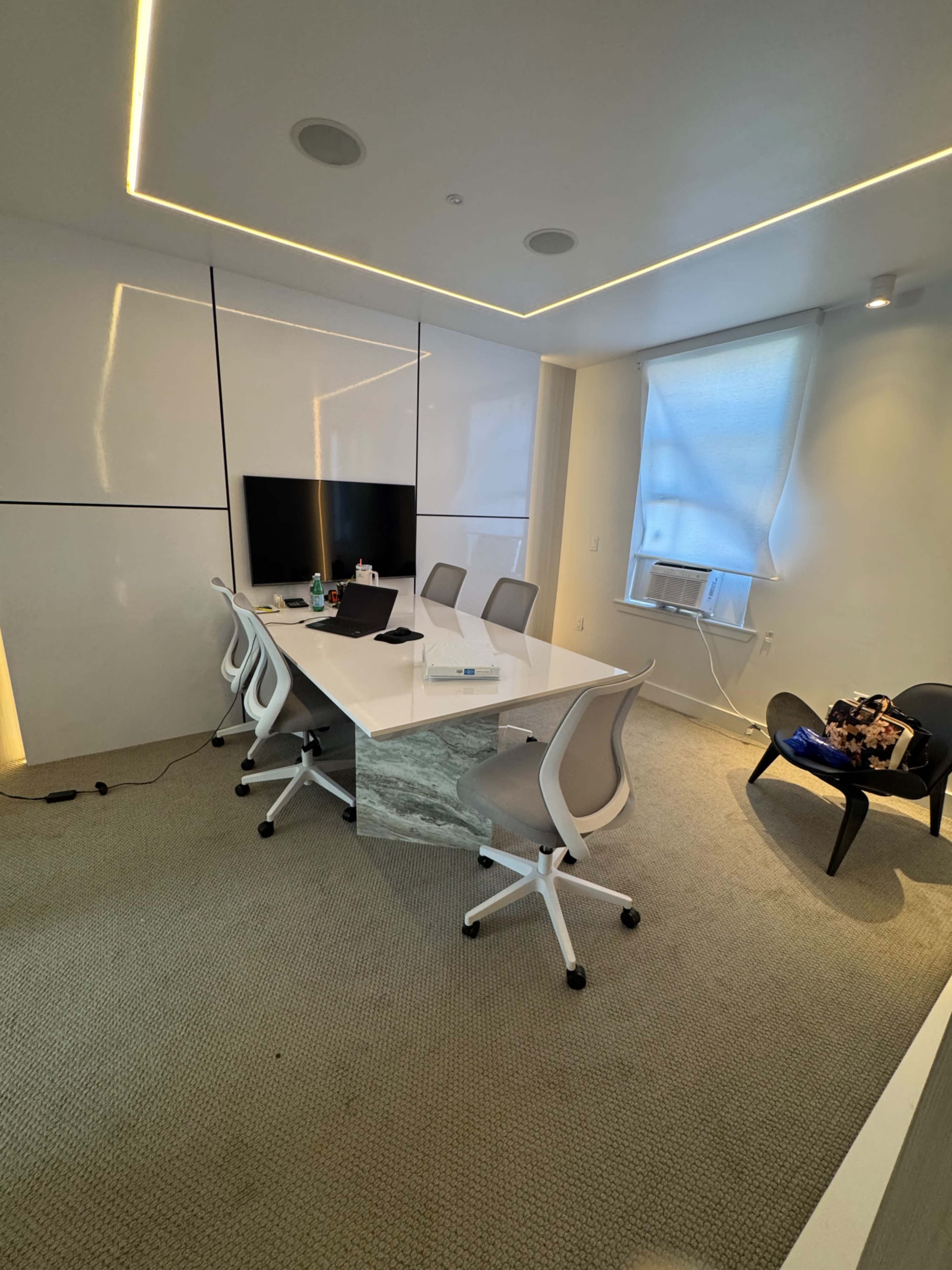 A modern conference room features a white table and chairs, a wall-mounted television, and a window with an air conditioning unit.