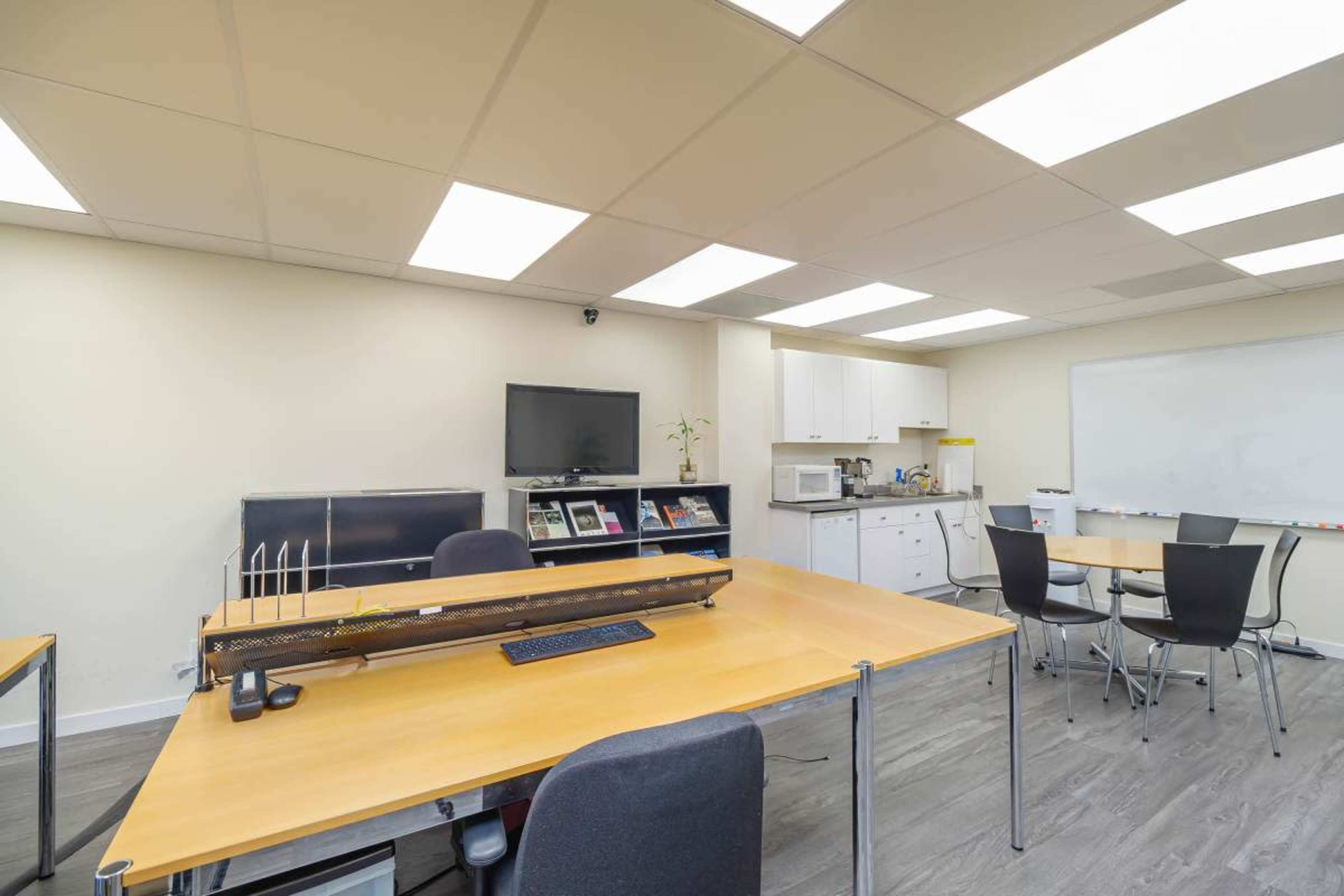 The image shows a modern office space with a large central table, chairs around it, a TV on the wall, and a kitchenette area.