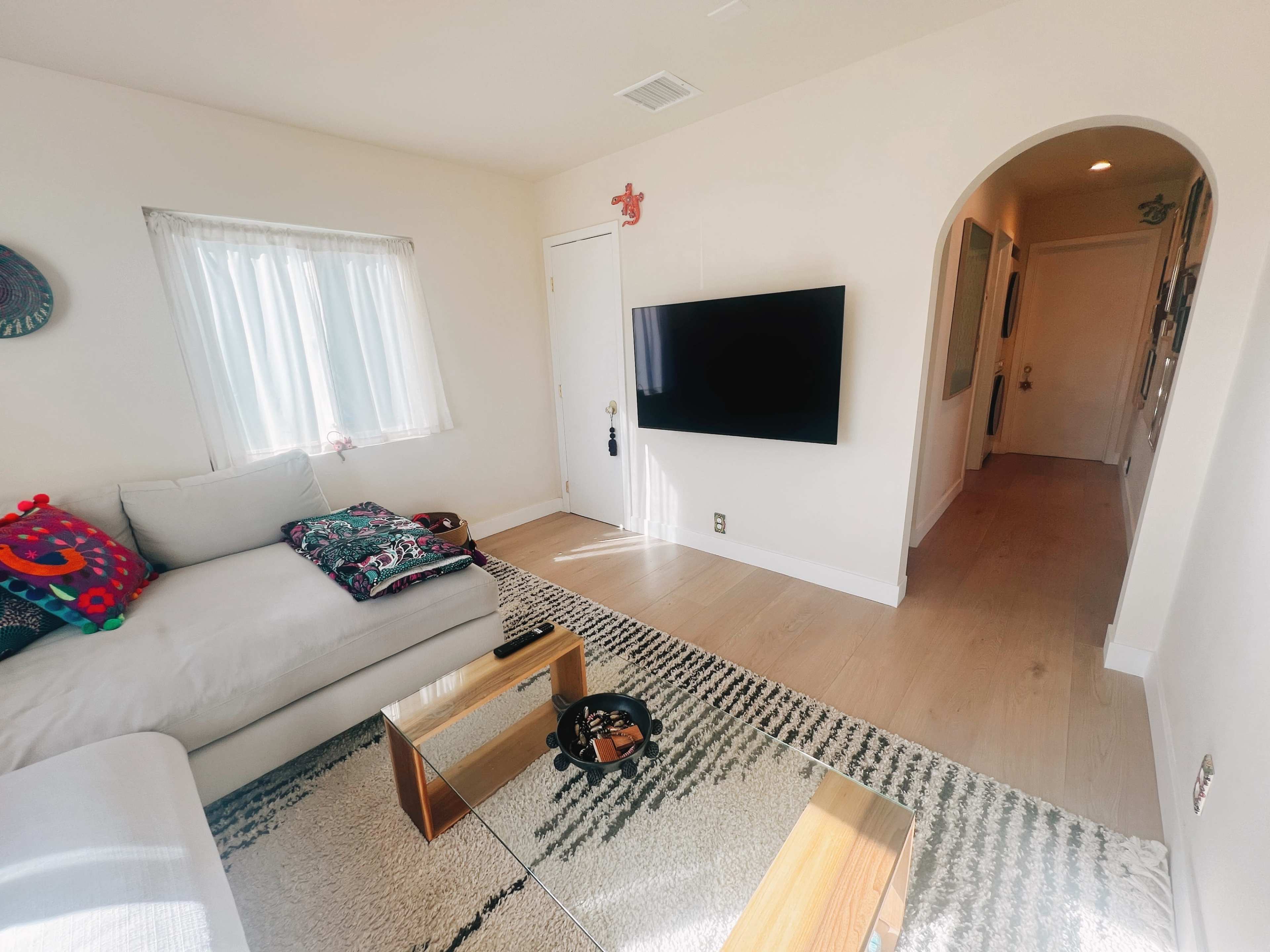 The image shows a bright living room with a light-colored sofa, a small coffee table, a mounted television, and an arched doorway leading to a hallway.
