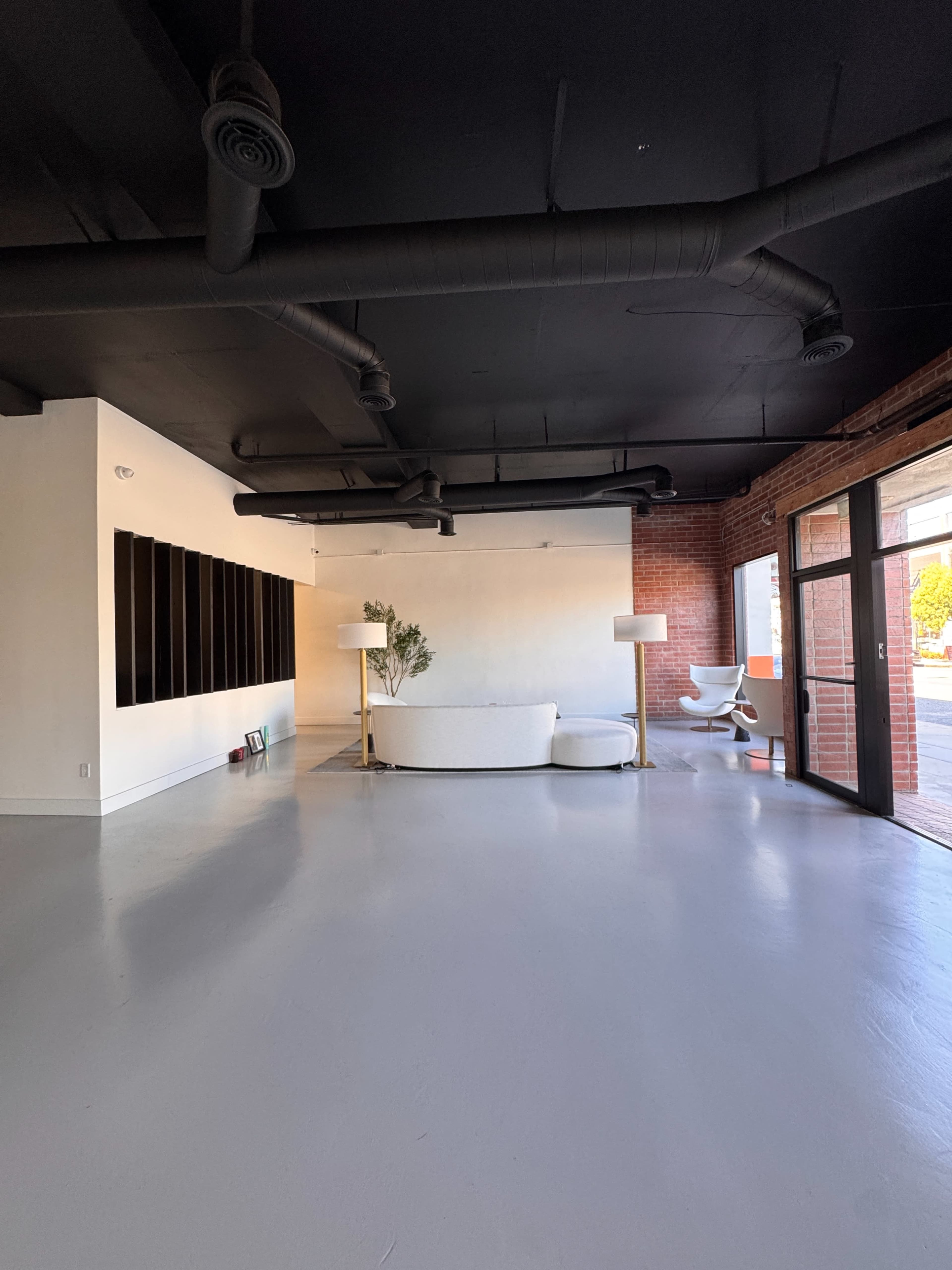 The image shows a spacious, modern interior with a white couch, a plant, a lamp, and large windows revealing an outdoor area.