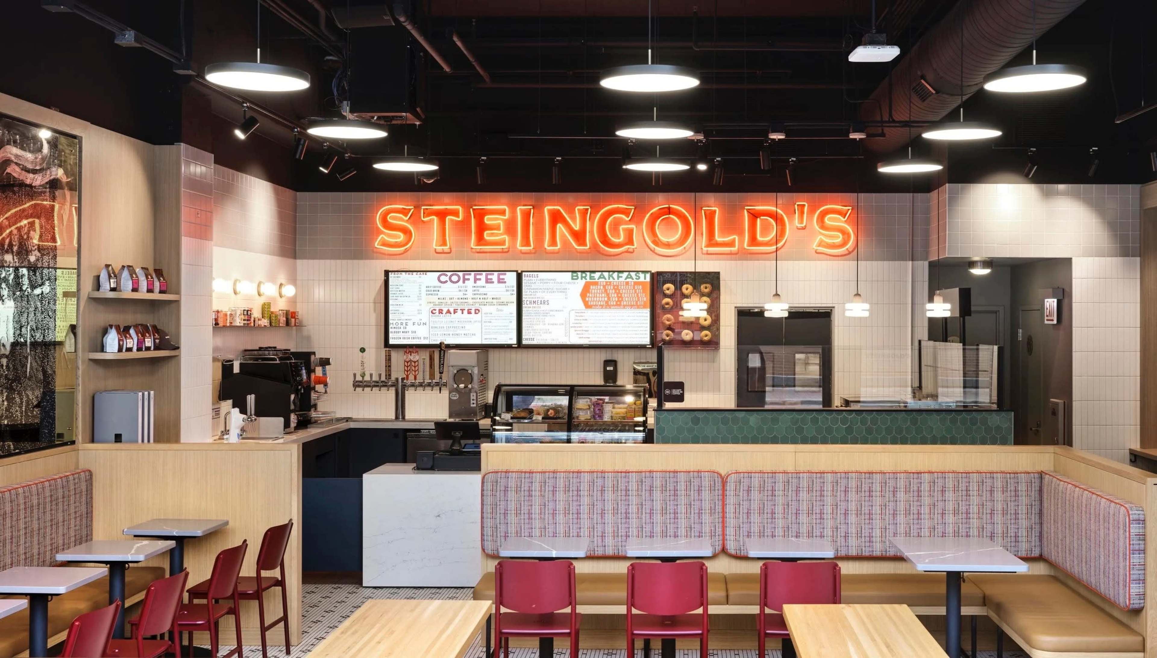 The image shows the interior of a modern café named "Steingold's," featuring a counter with a menu, seating areas with tables and red chairs, and neon signage.