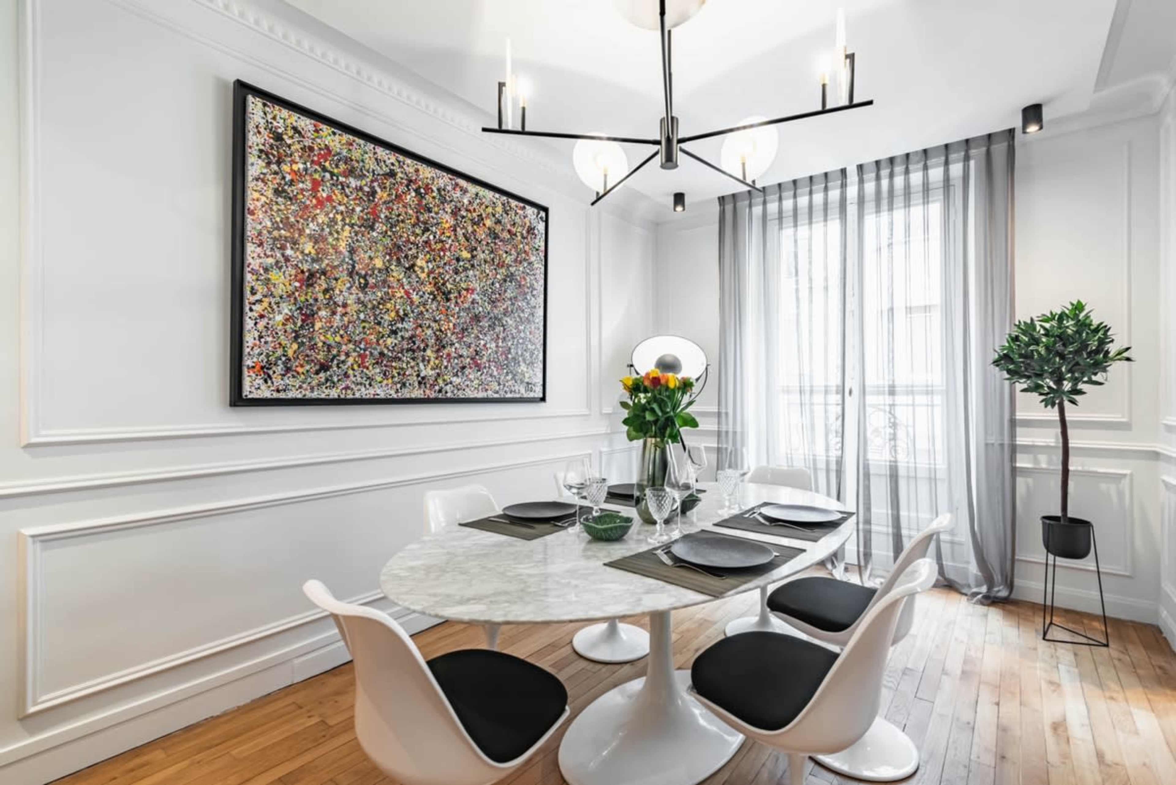 A modern dining room features a round marble table set for four, with a large colorful abstract painting on the wall and a bouquet of flowers in the center.