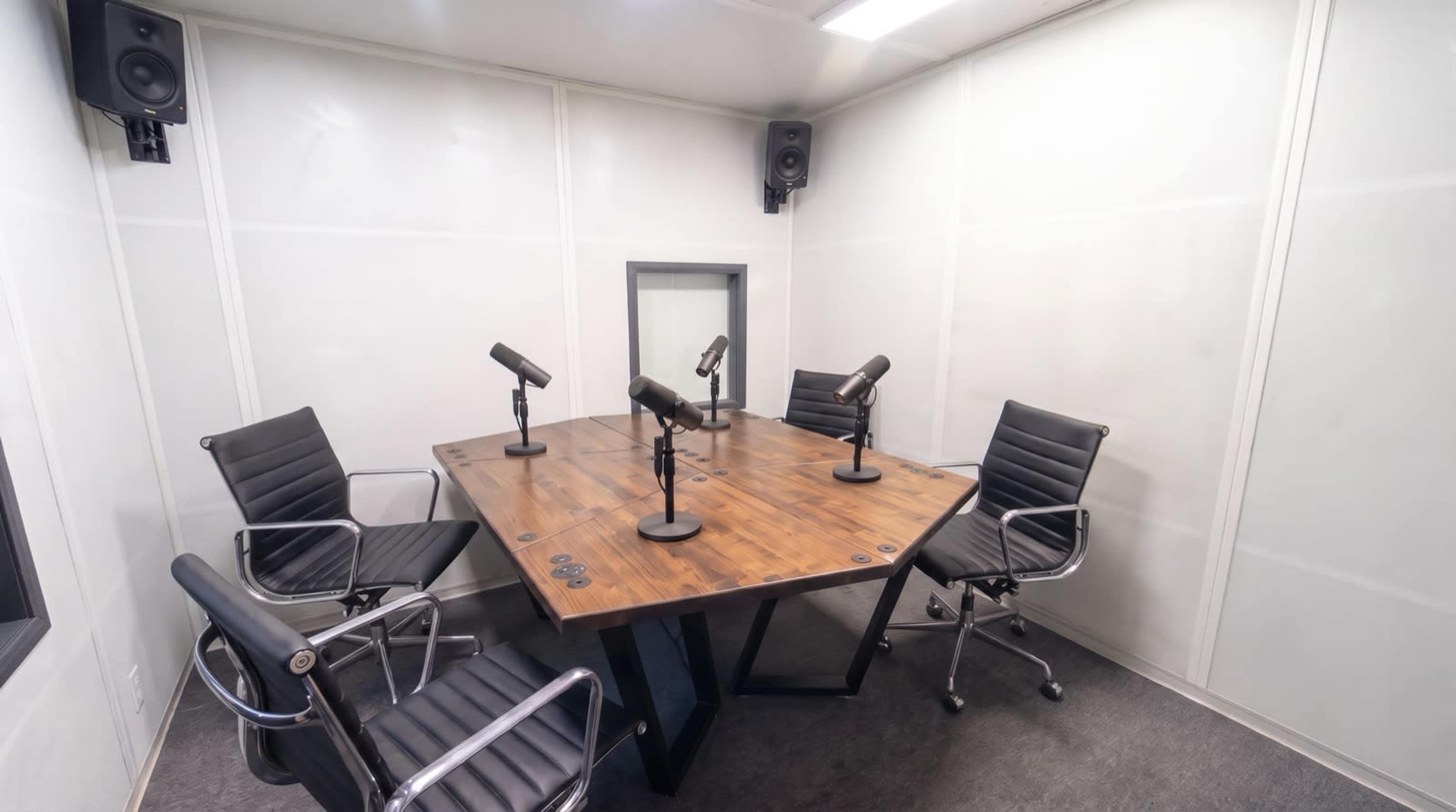 The image shows a small recording studio equipped with a wooden table, four black chairs, and microphones positioned for podcasting or interviews.