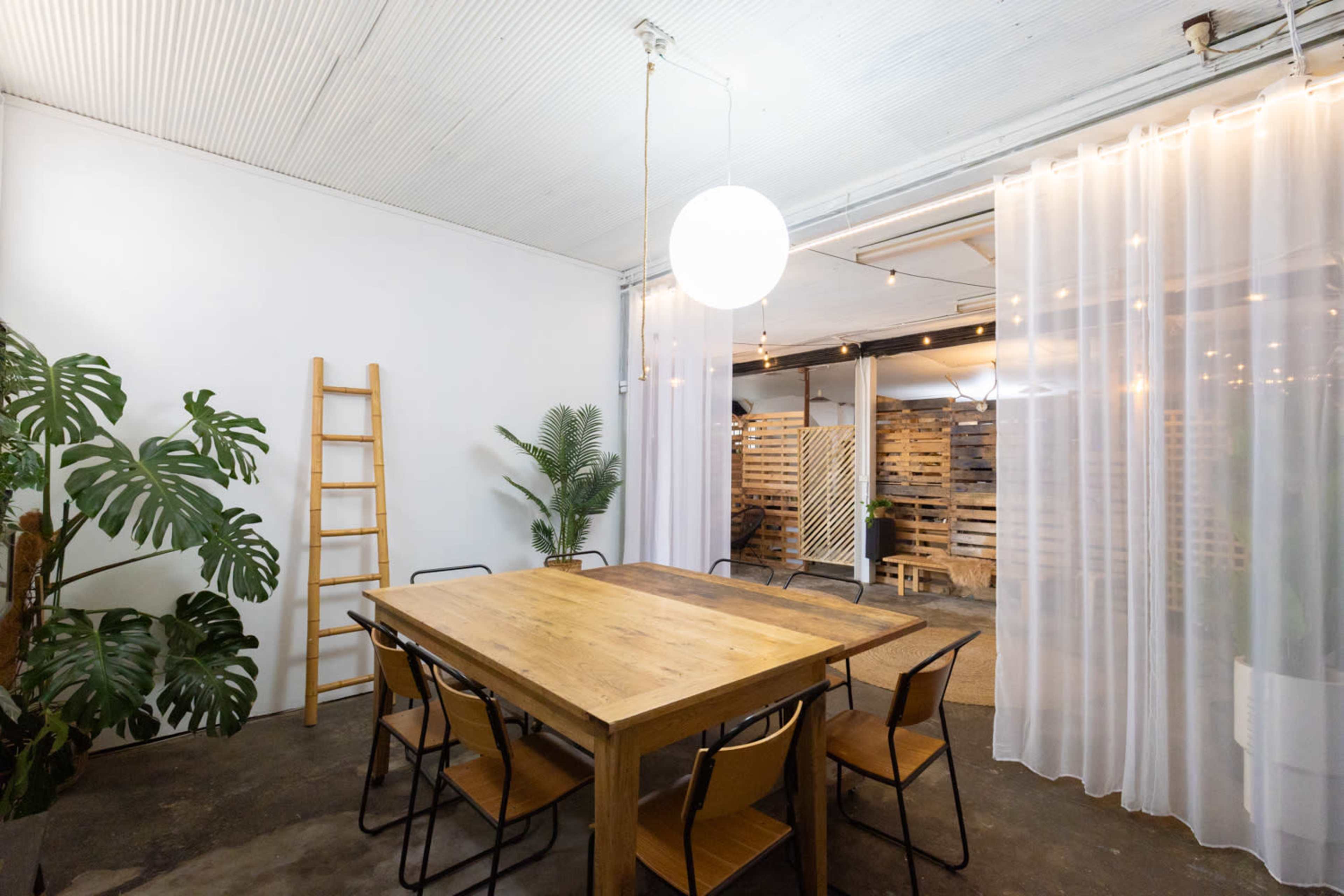 A wooden dining table surrounded by black chairs is located in a minimalist room with white walls and a hanging light fixture, with a curtain and ladder visible in the background.
