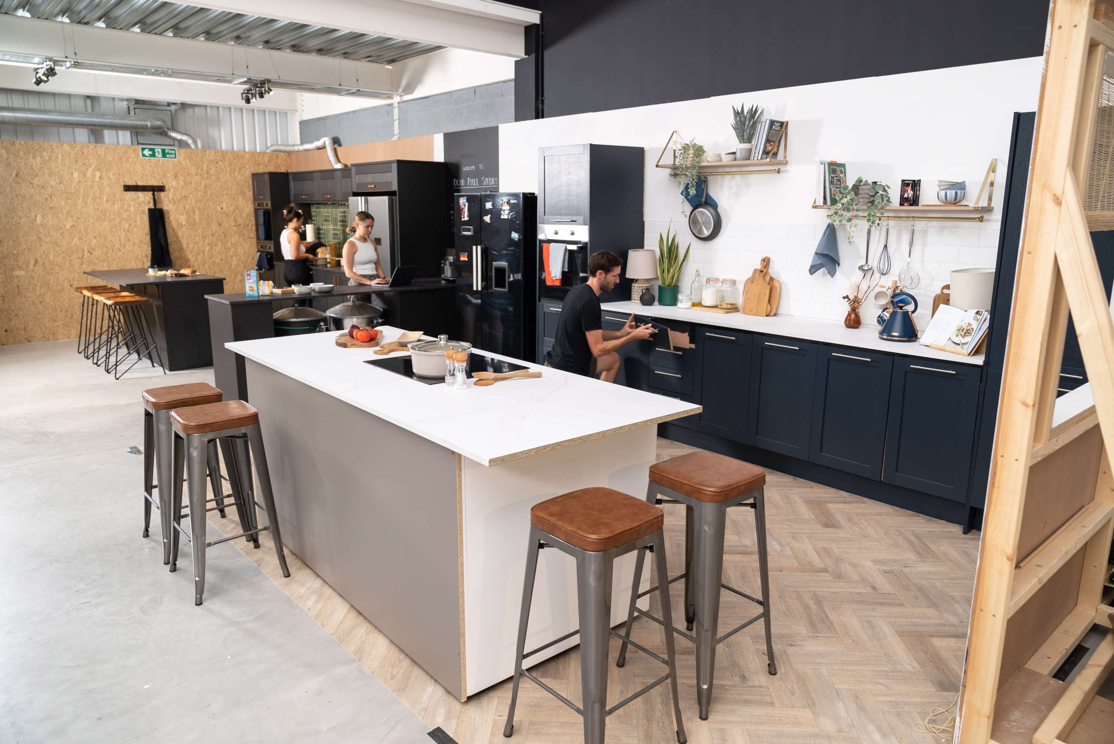 A modern kitchen features a central island with seating, black cabinetry, and a person preparing food.