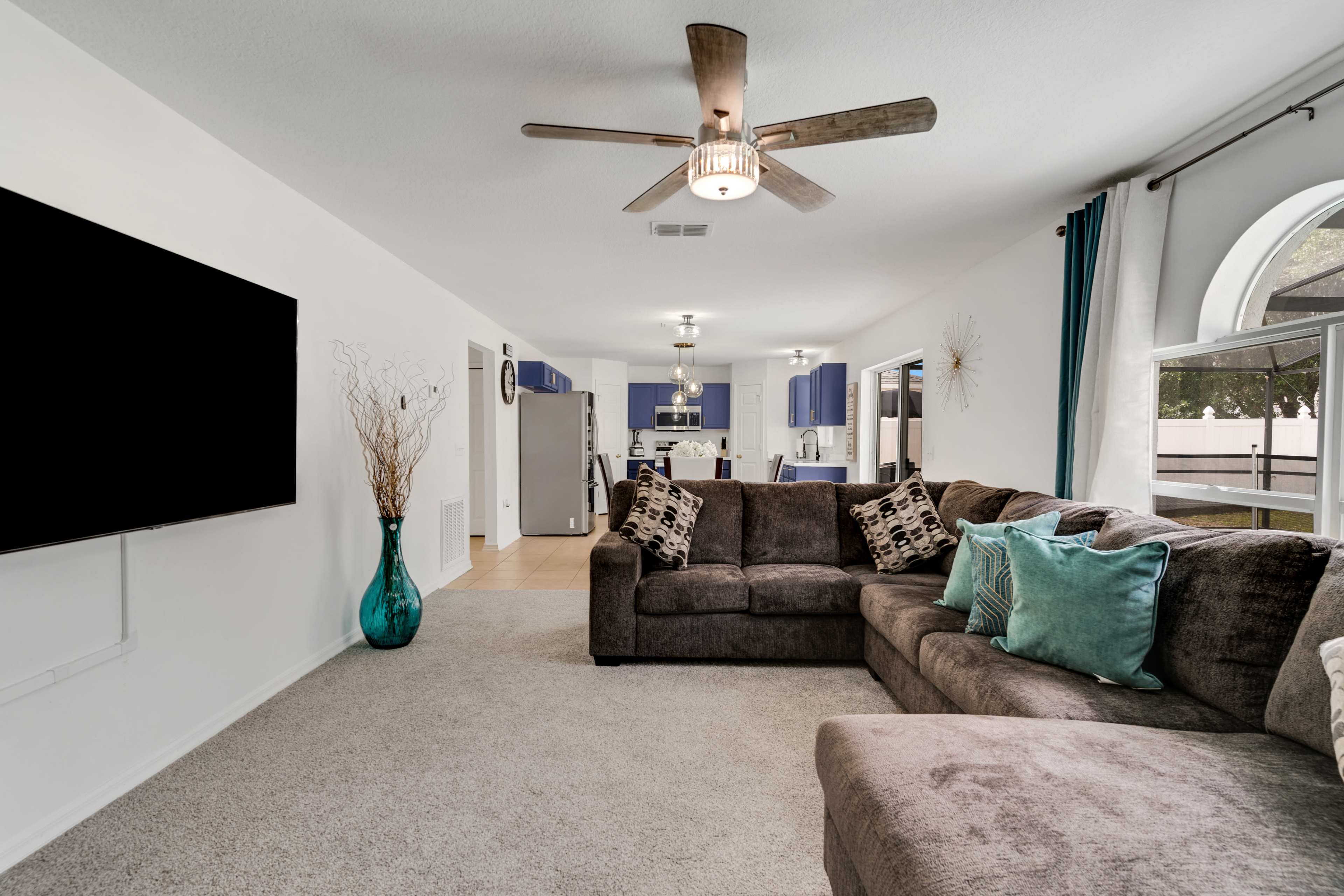 A spacious living room features a large sectional couch, a television mounted on the wall, and a view of a kitchen in the background.