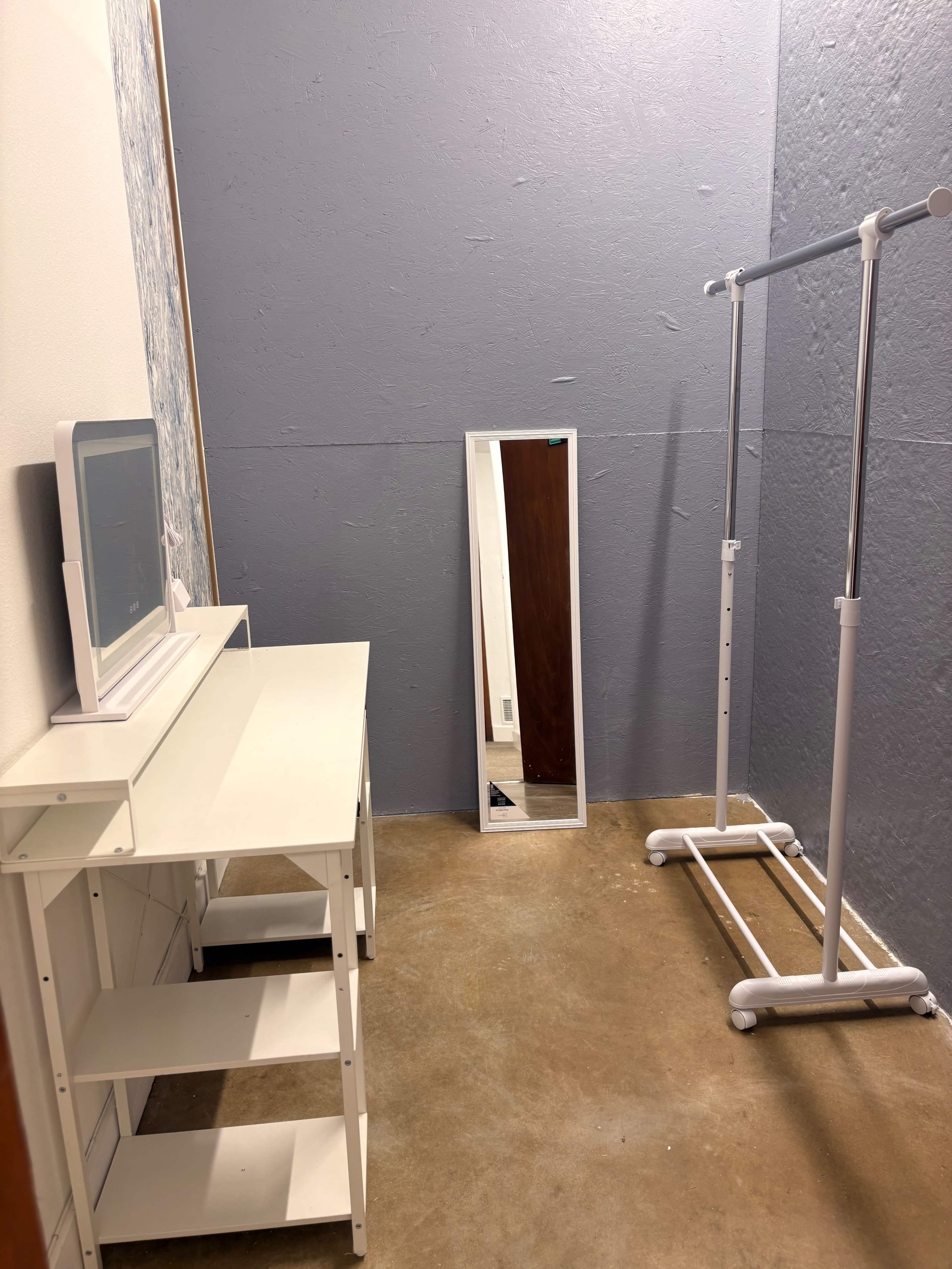 The image shows a small room with a gray wall, a white desk with shelves, a full-length mirror, and a clothing rack.