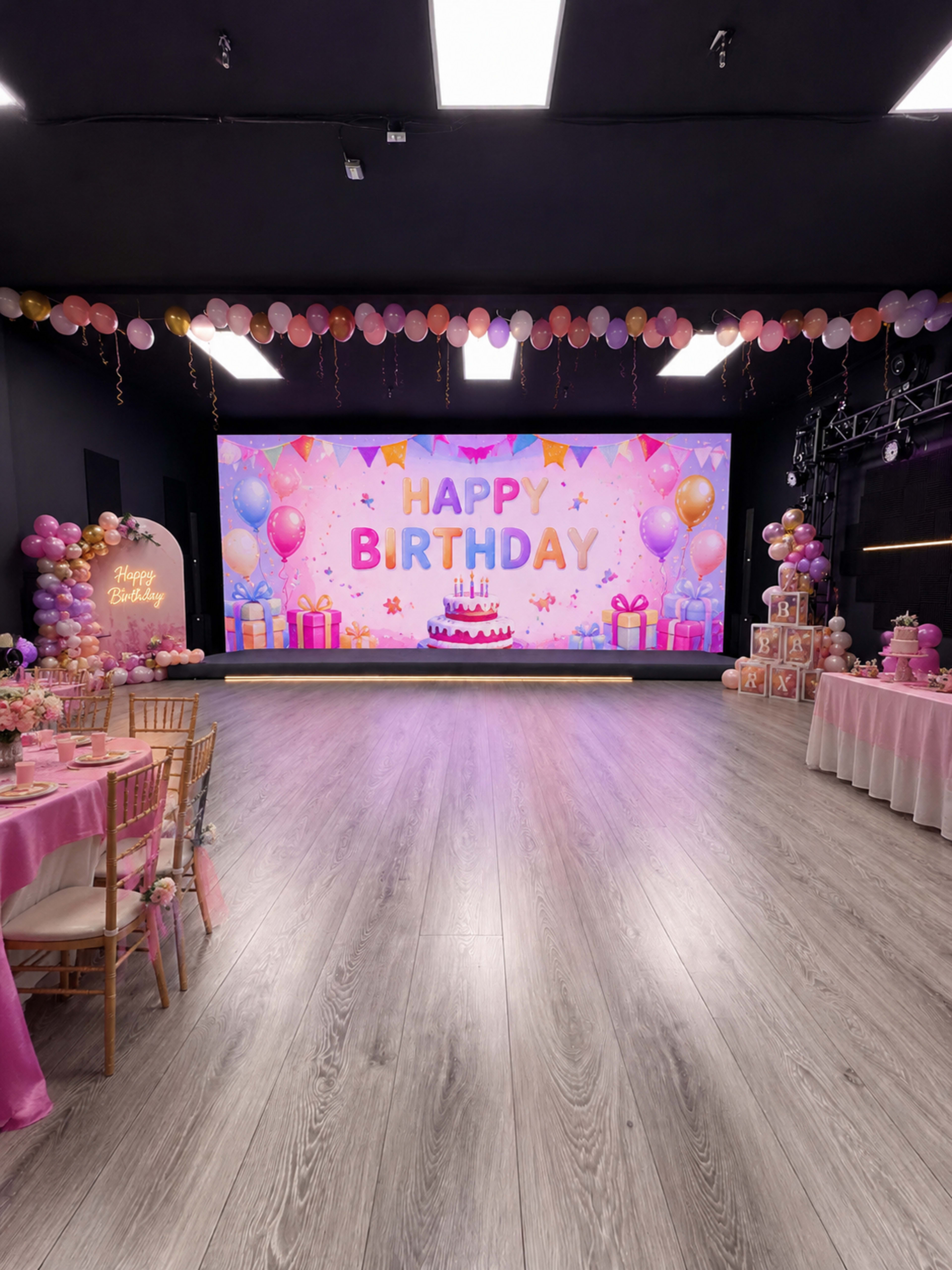 The image shows a decorated indoor venue for a birthday celebration, featuring a large screen displaying "Happy Birthday" and festive balloons and tables set up for guests.