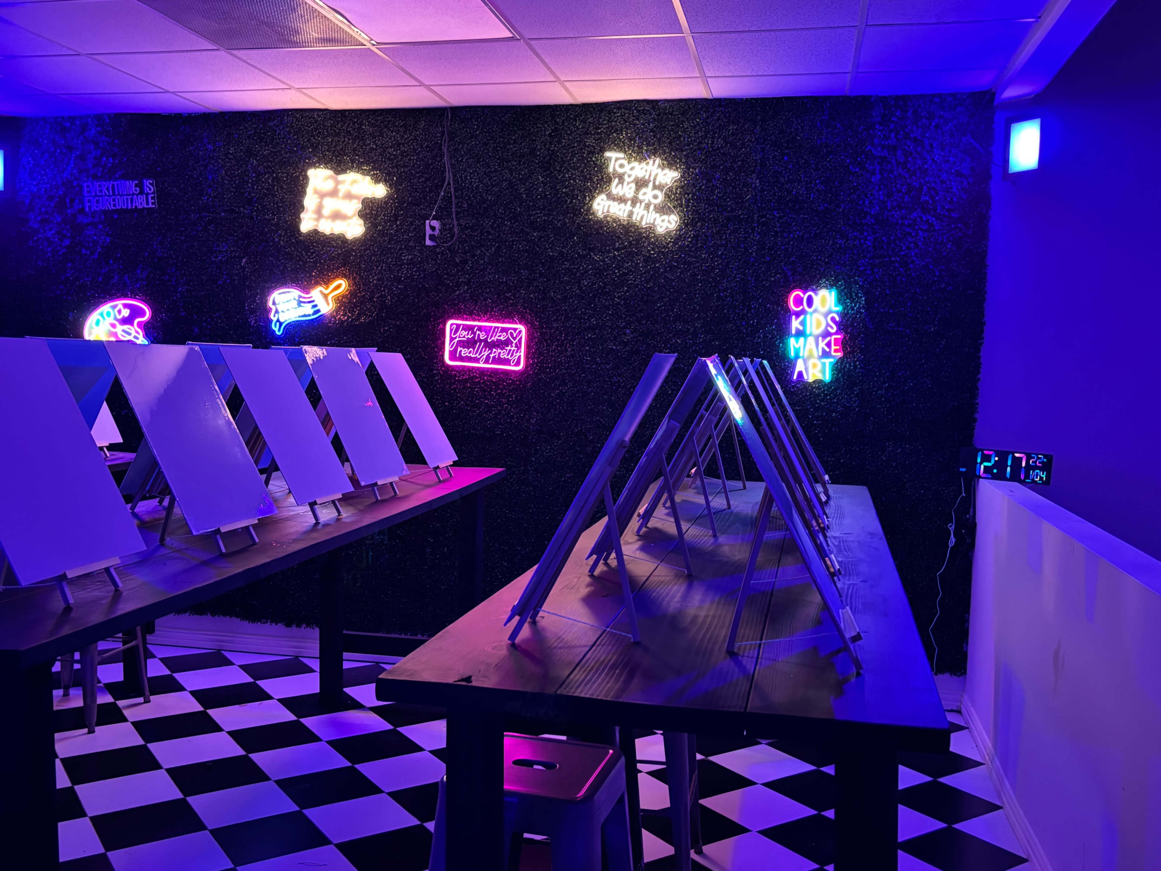 The image shows an art studio featuring easels arranged on tables with neon signs and a black-and-white checkered floor.