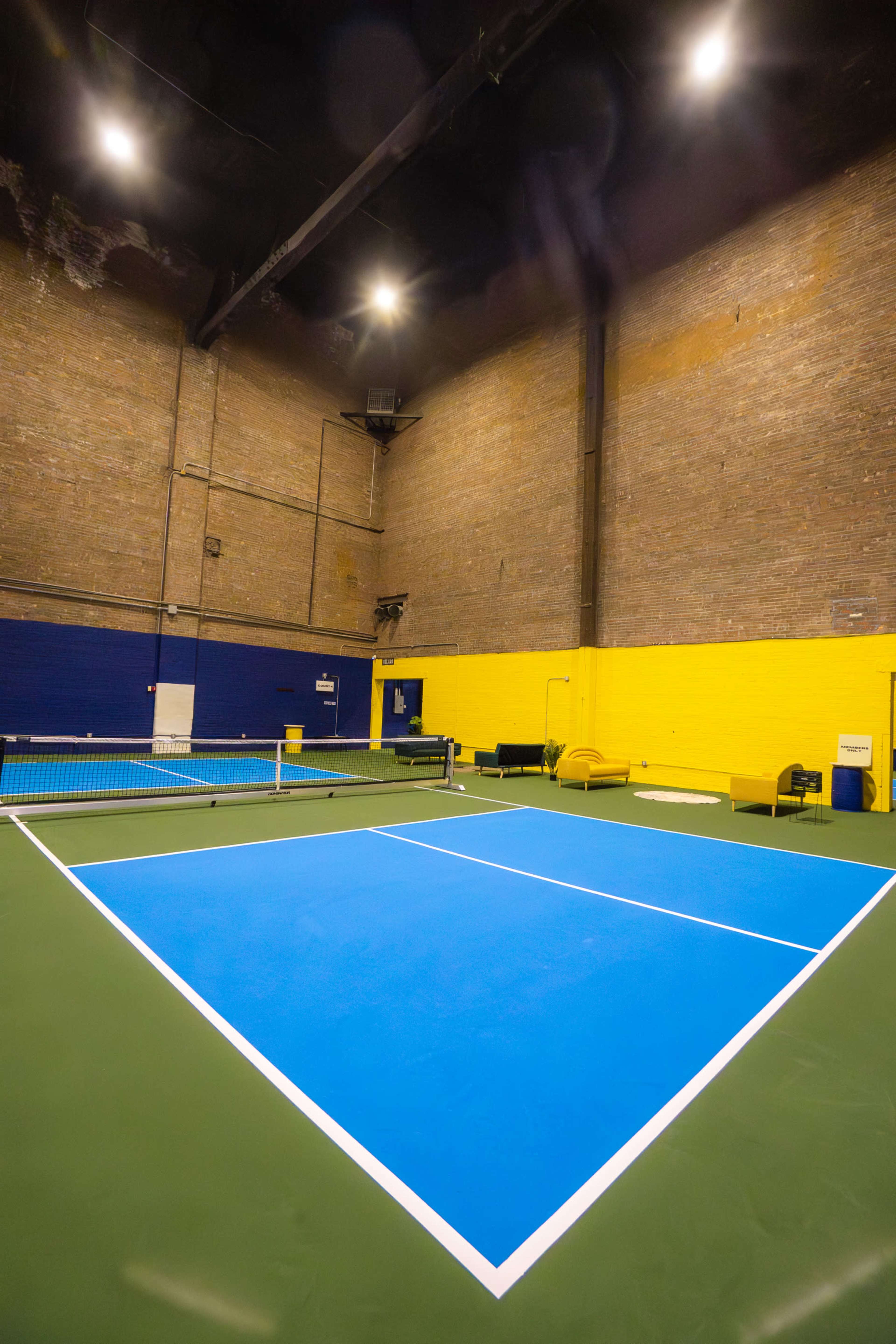 The image shows an indoor tennis court with a blue playing surface, green perimeter, and yellow and blue walls.
