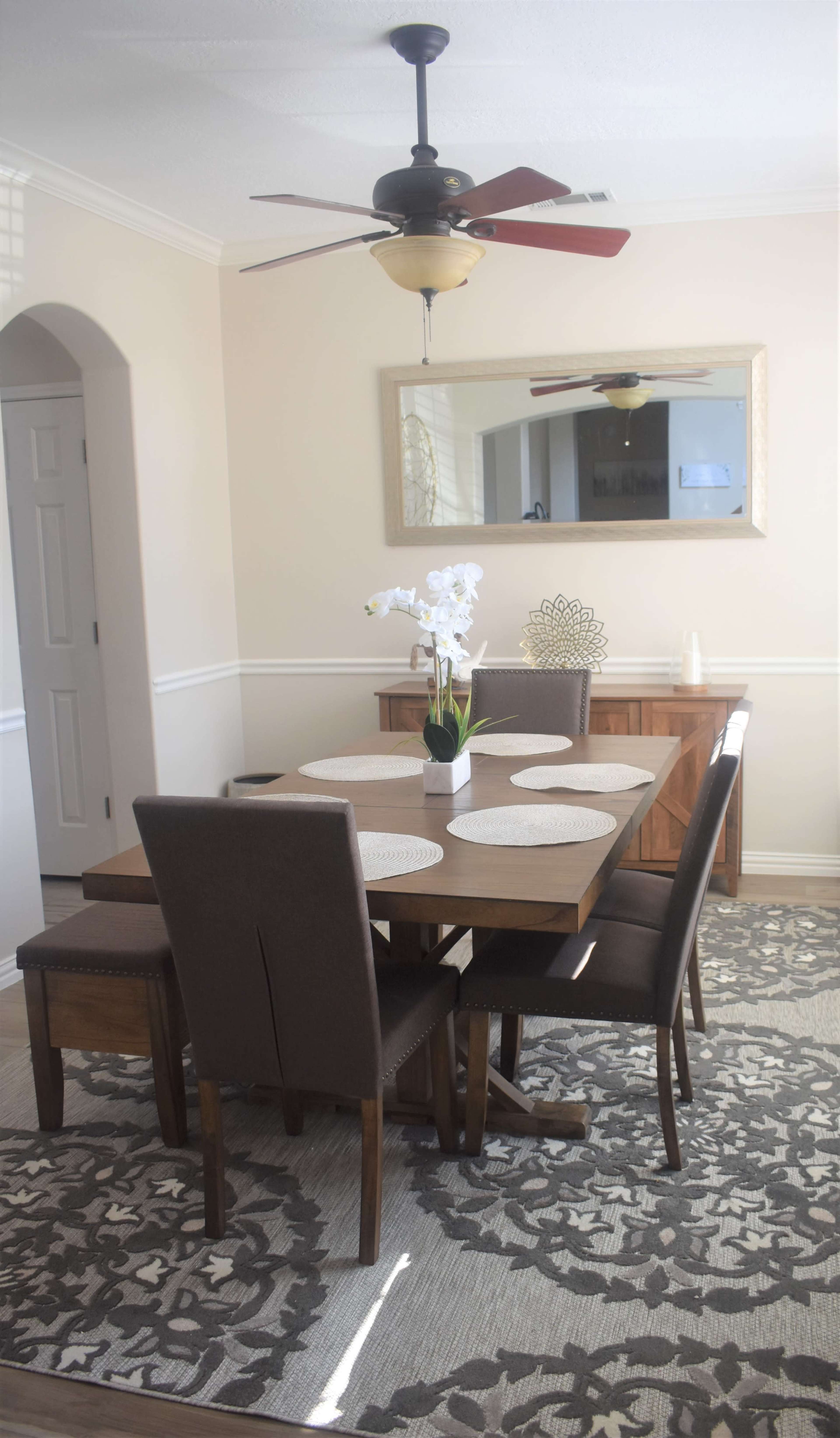 A dining room with a wooden table surrounded by four chairs, a mirror on the wall, and decorative elements including a vase of flowers on the table.