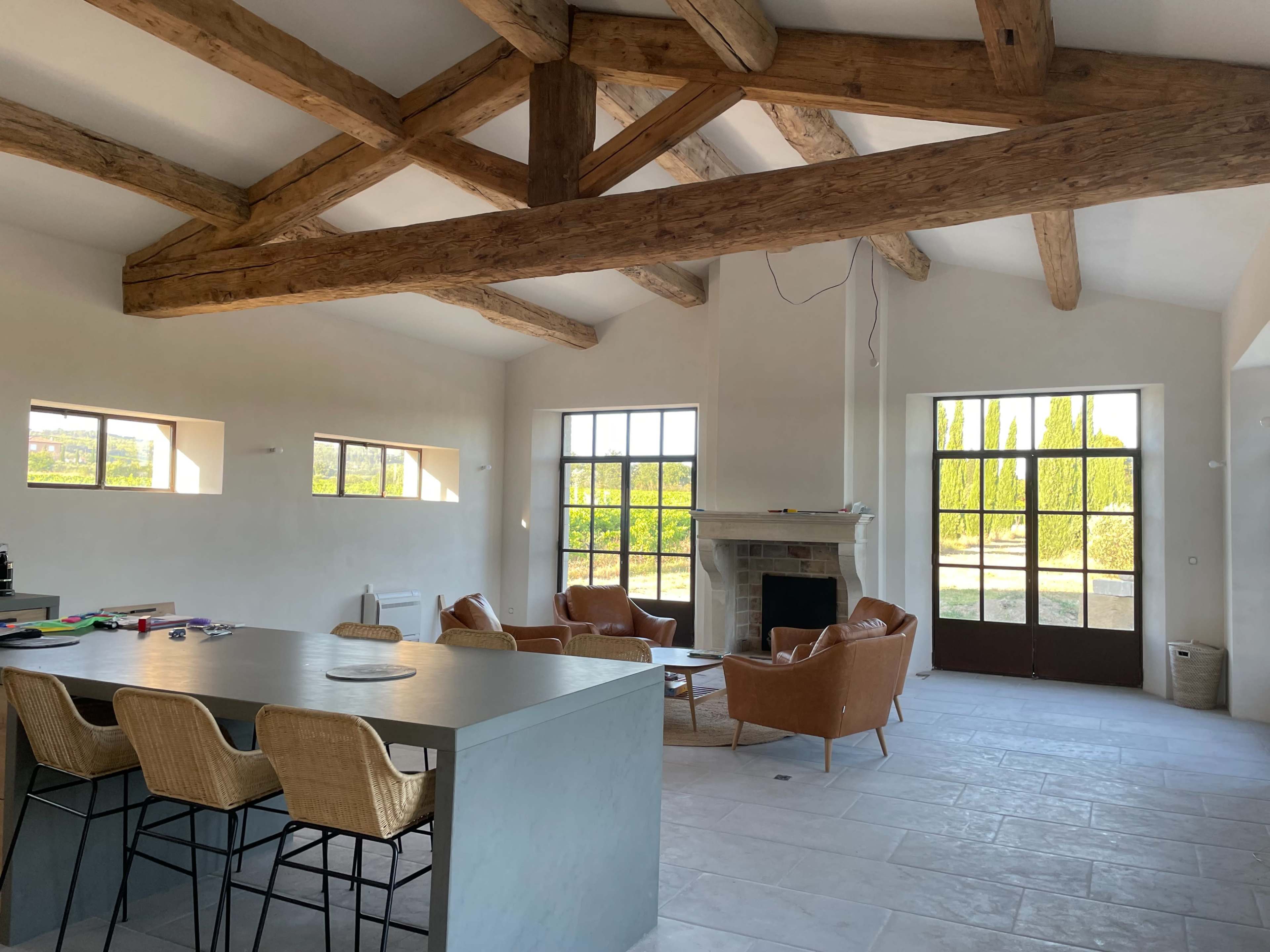 The image shows an open-concept living space with exposed wooden beams, a stone fireplace, and large windows overlooking a green landscape.