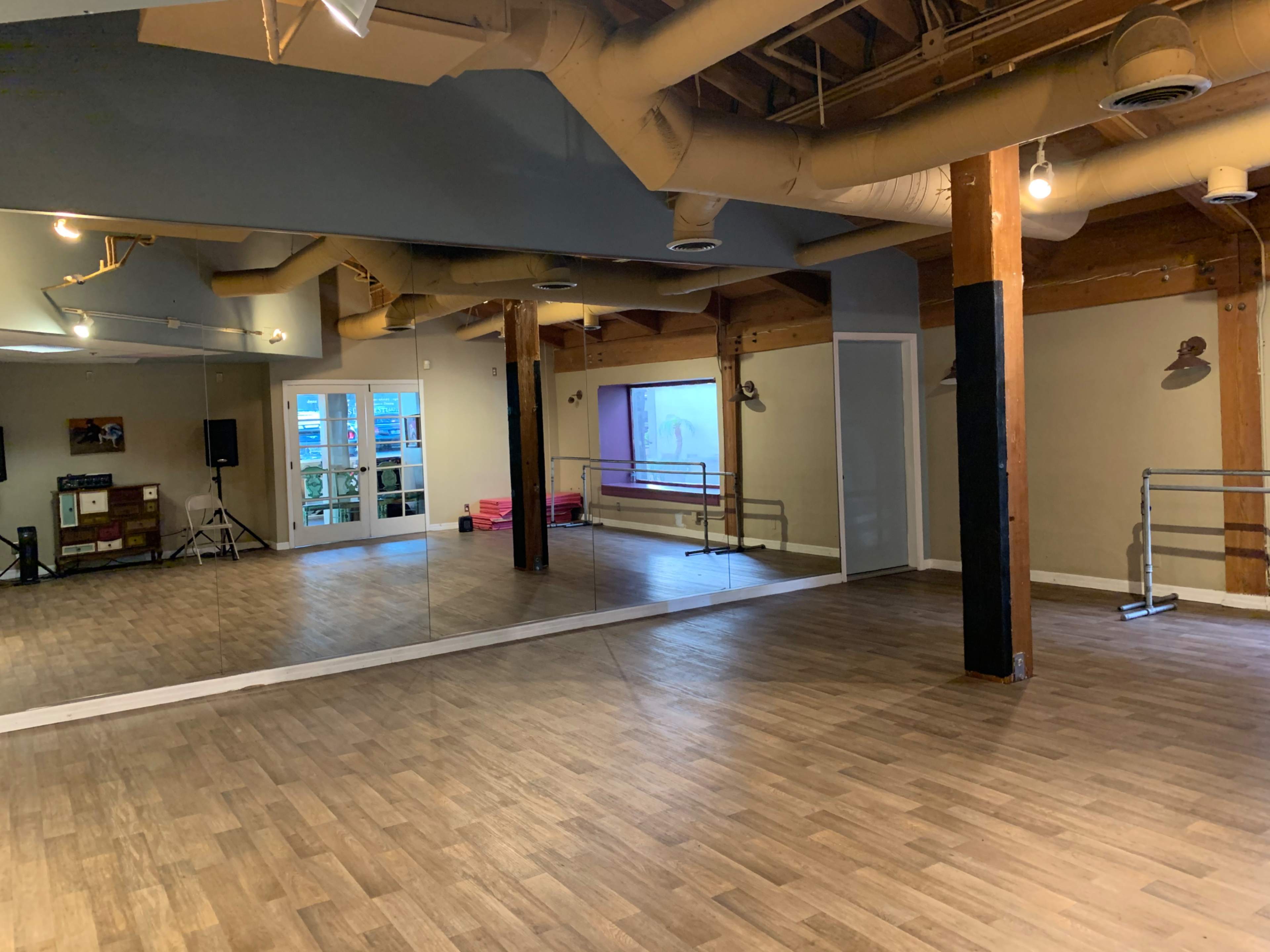 An empty dance studio with wooden flooring, mirrors on one wall, and large windows that allow natural light to enter.