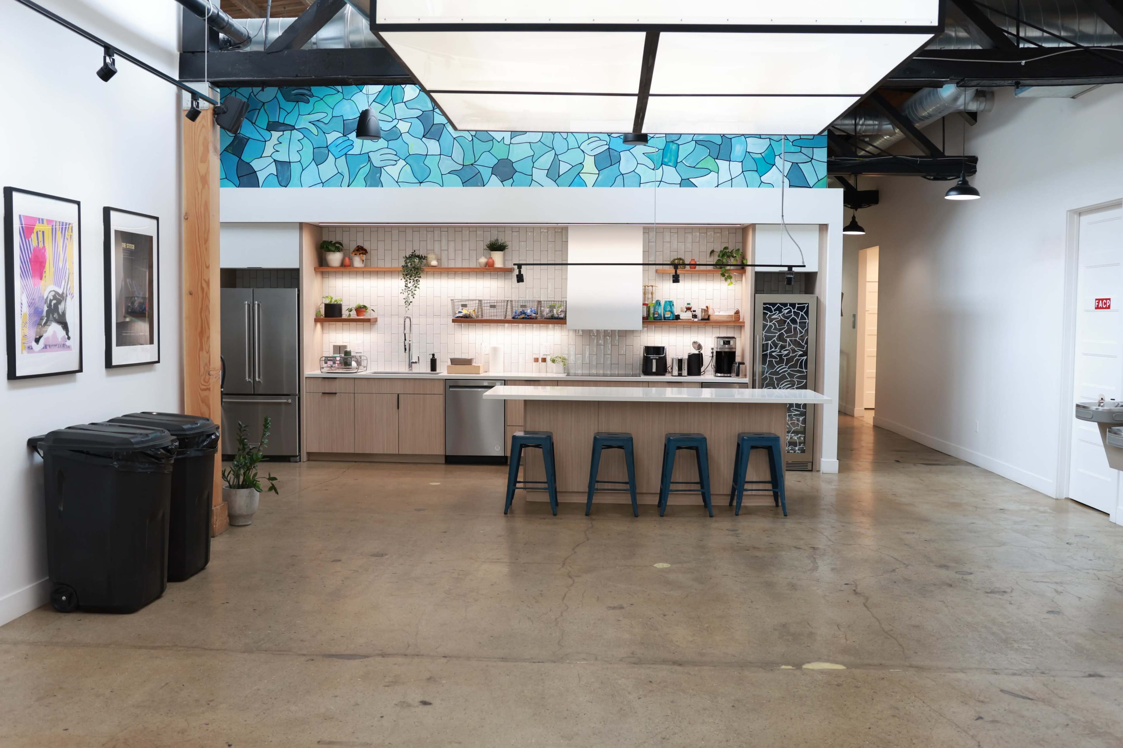 The image shows a modern kitchen area with a central island, bar stools, and sleek cabinetry, set against a backdrop of colorful wall art and plants.
