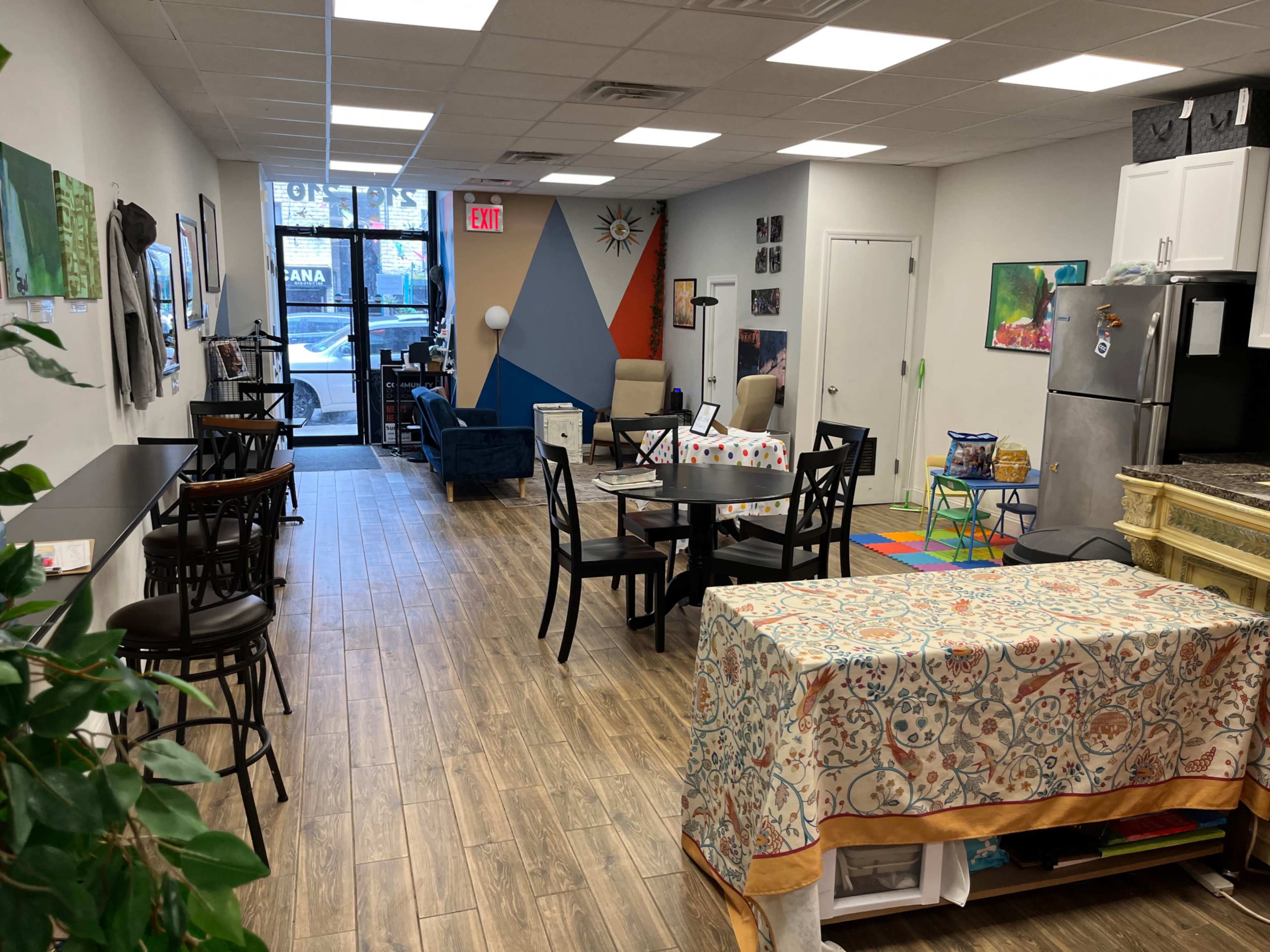 The image features a brightly lit communal space with multiple seating areas, including tables and chairs, colorful decor, and a kitchenette.