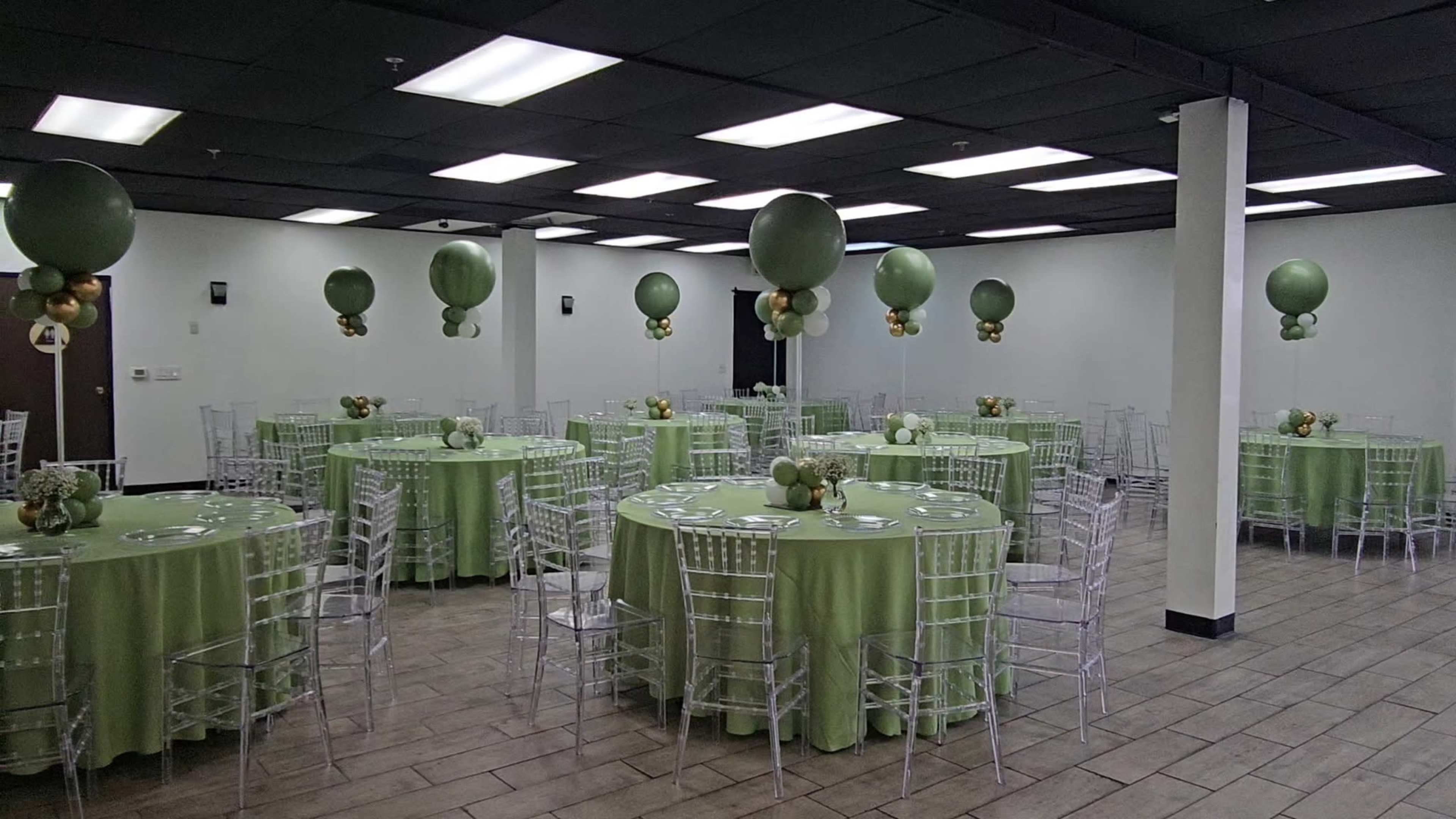 The image shows a banquet hall with multiple round tables covered in light green tablecloths, each adorned with centerpieces and surrounded by transparent chiavari chairs.