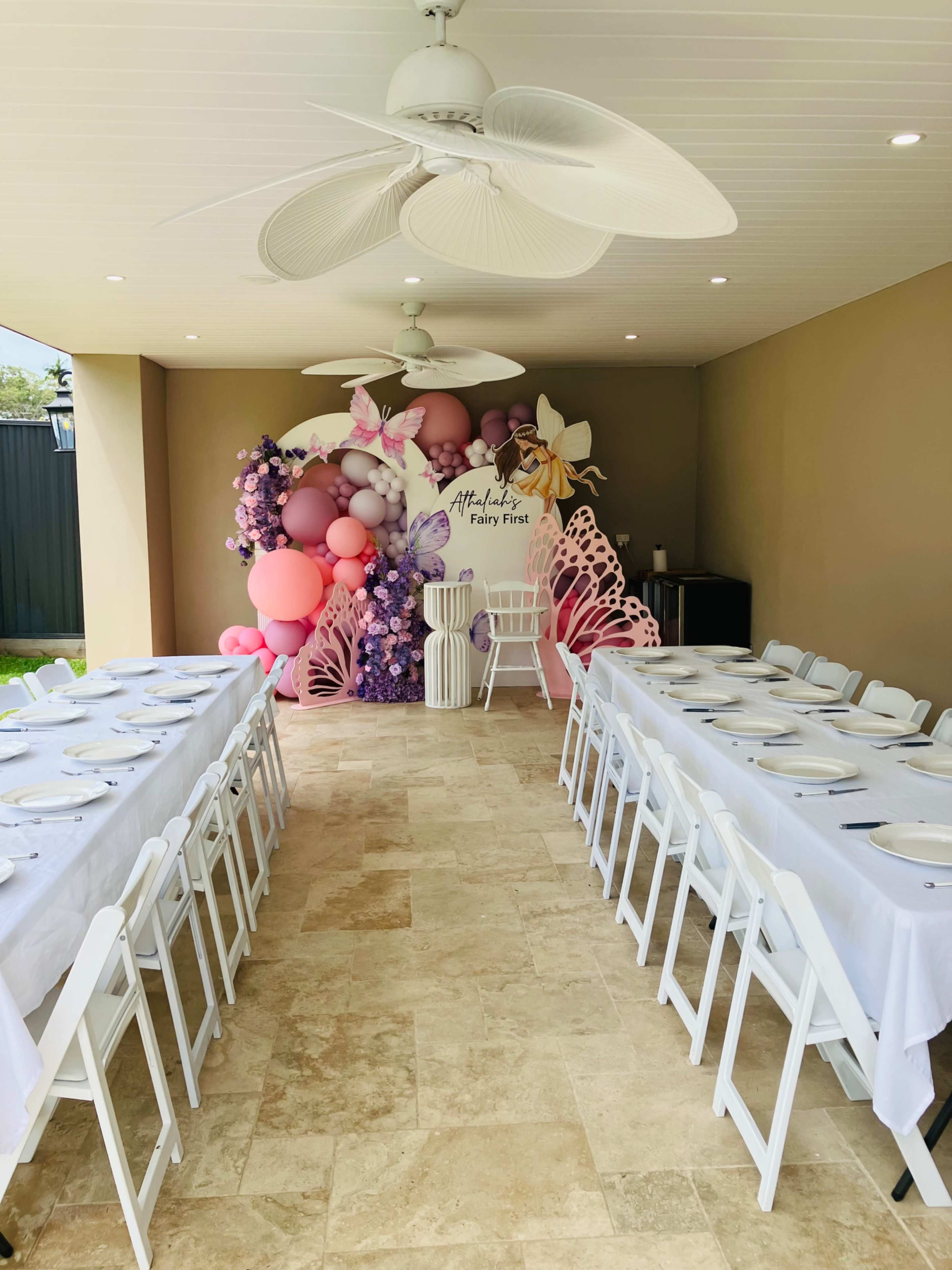 The image shows a decorated event space featuring a long white table set with plates, surrounded by white chairs, and a colorful balloon display in the background.