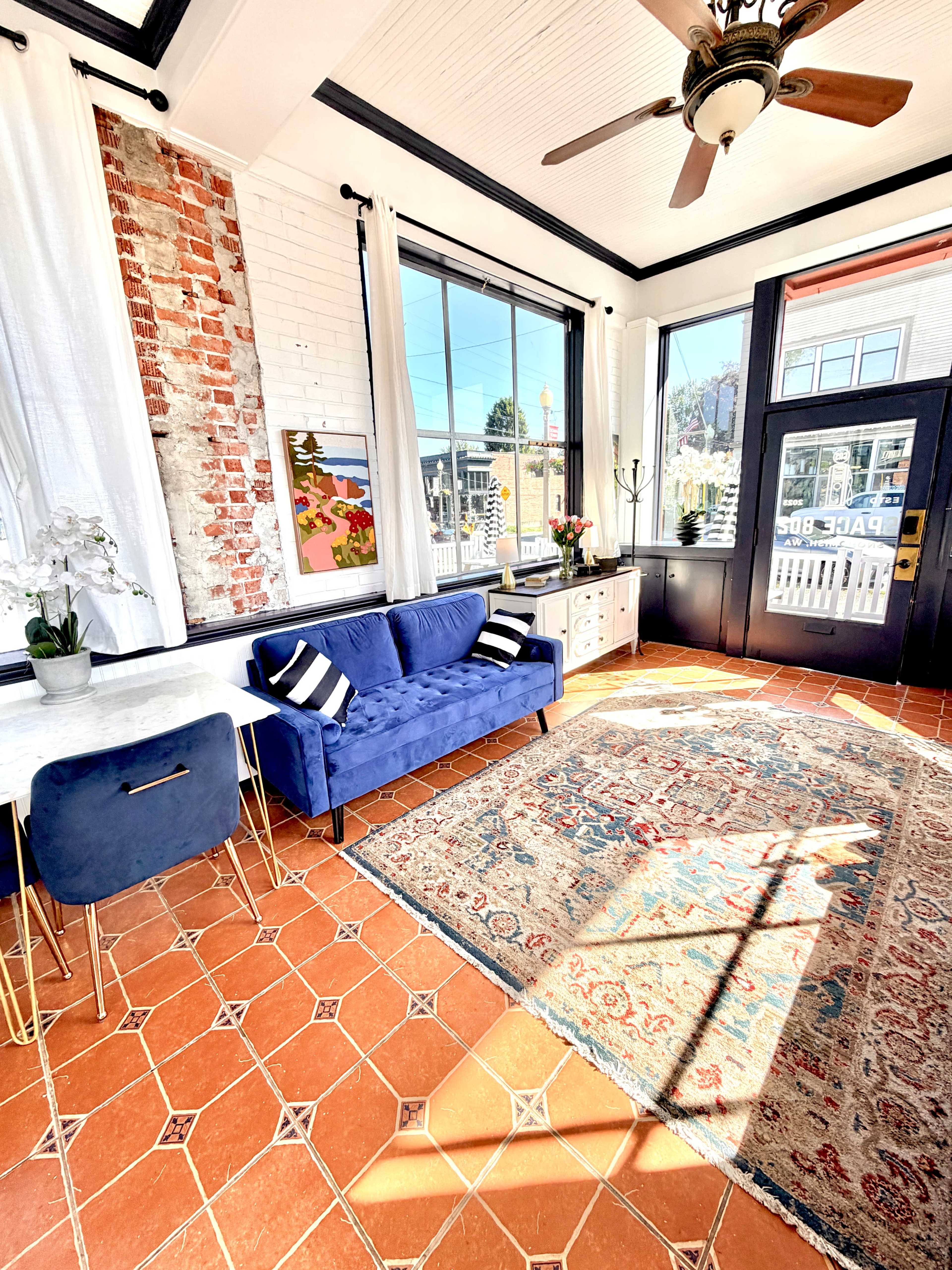 The image shows a well-lit interior space featuring a blue couch, a marble table, and a colorful rug on tiled flooring, with large windows allowing natural light to enter.