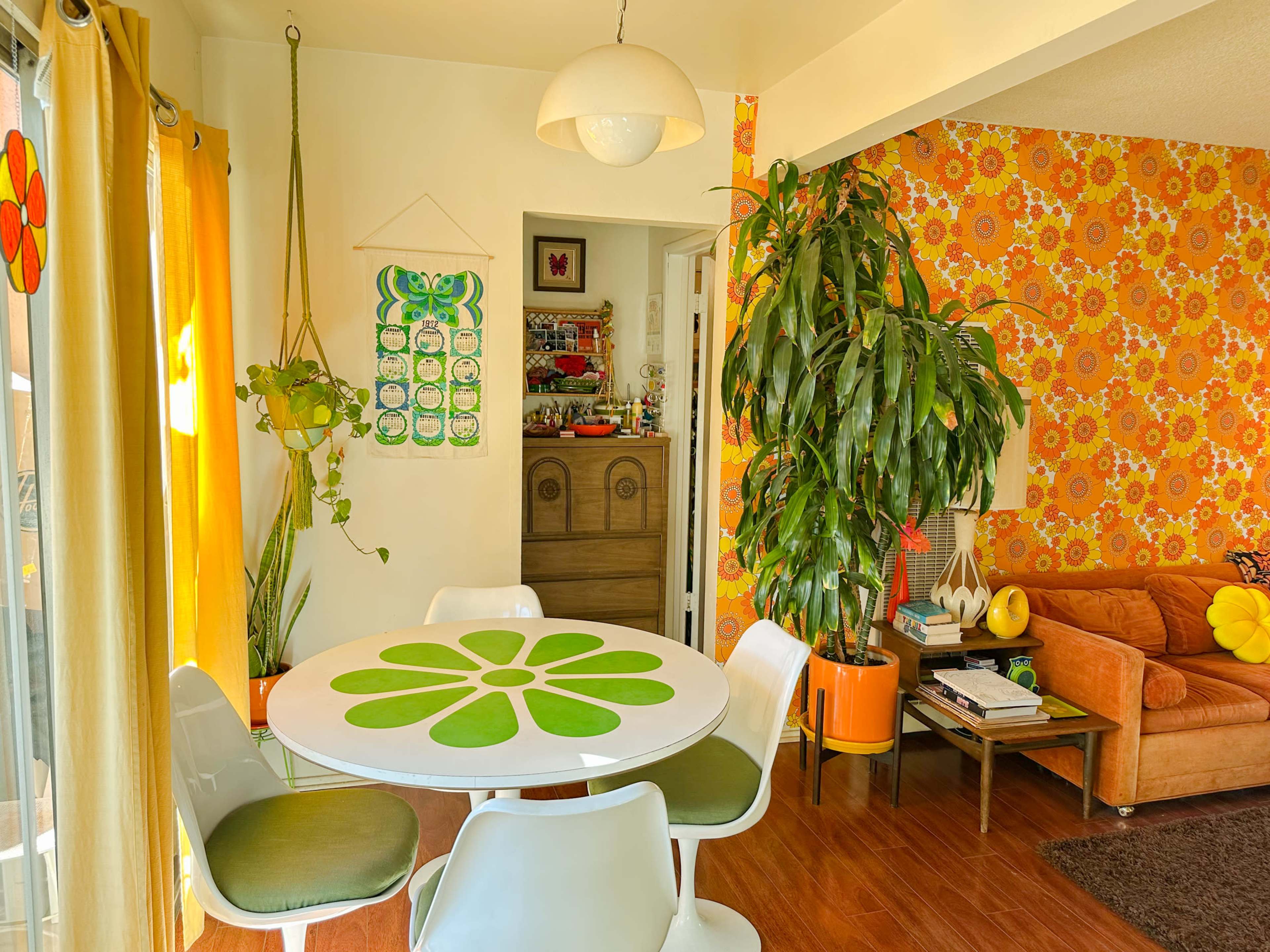 The image shows a brightly decorated dining area featuring a round table with a green floral design, white chairs, yellow curtains, and a wall adorned with orange floral wallpaper.