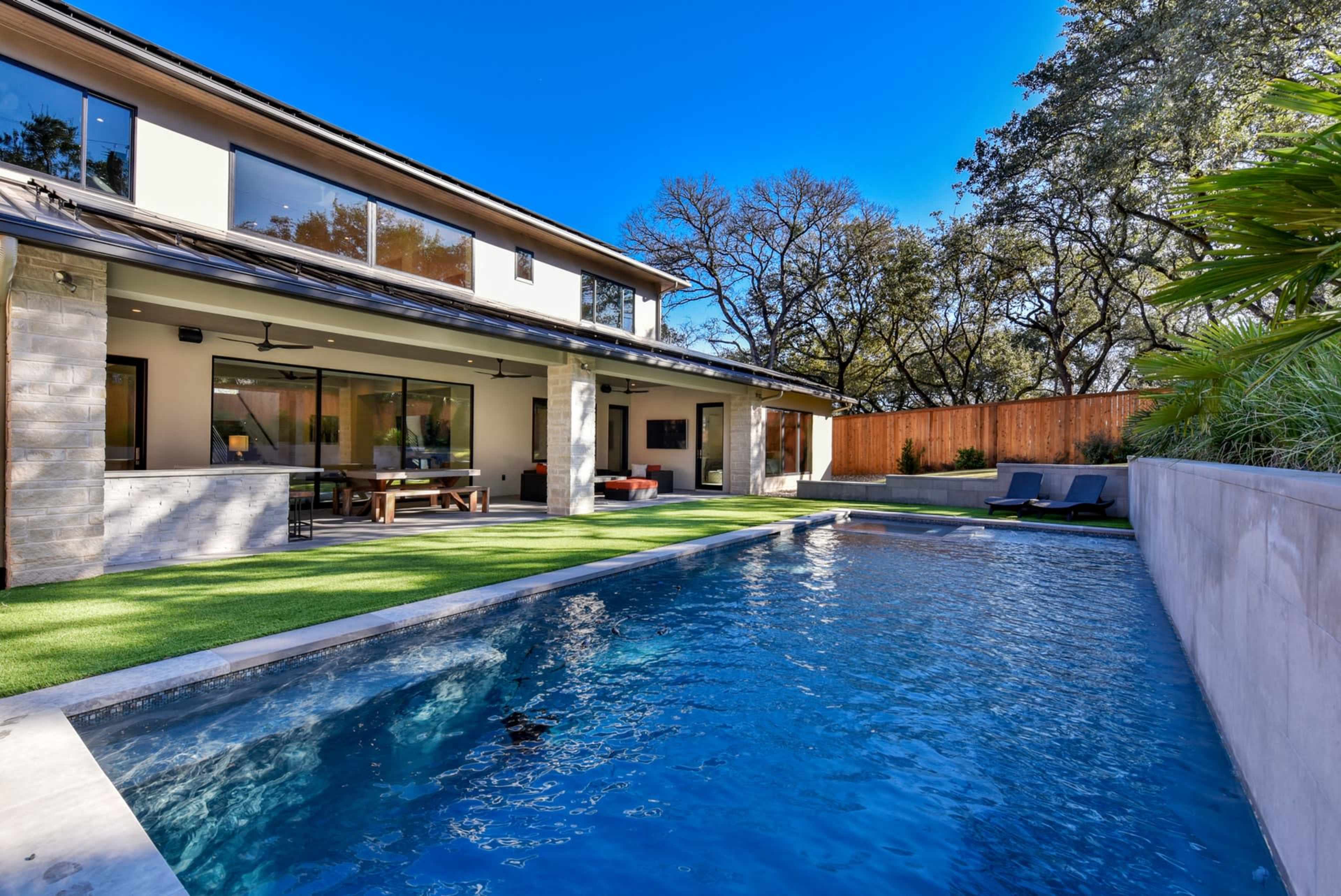 The image shows a modern two-story house with a clear blue pool in the backyard surrounded by green grass and trees.