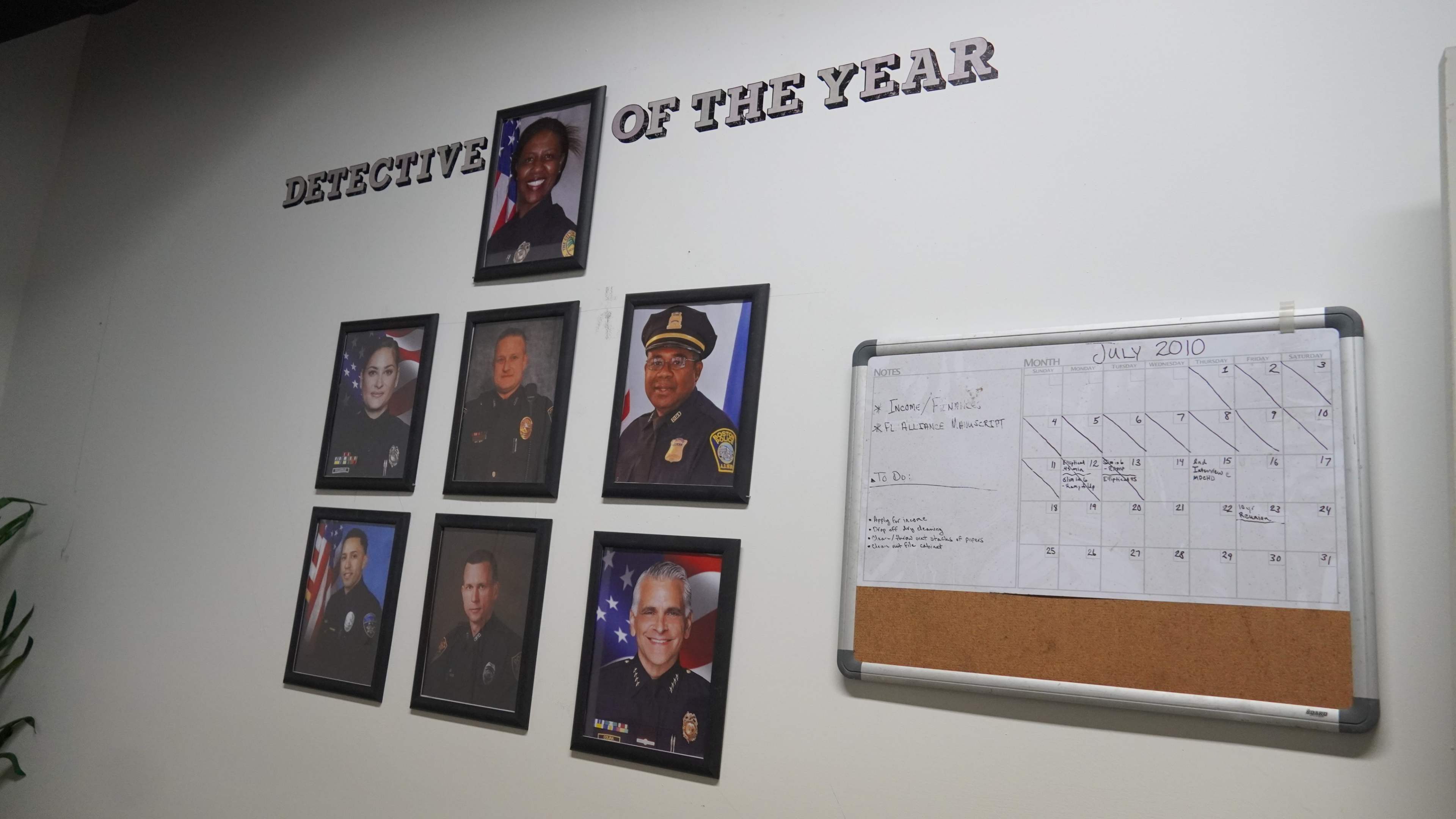 The image shows a wall display featuring framed portraits of several individuals recognized as "Detective of the Year," along with a calendar for July 2010.