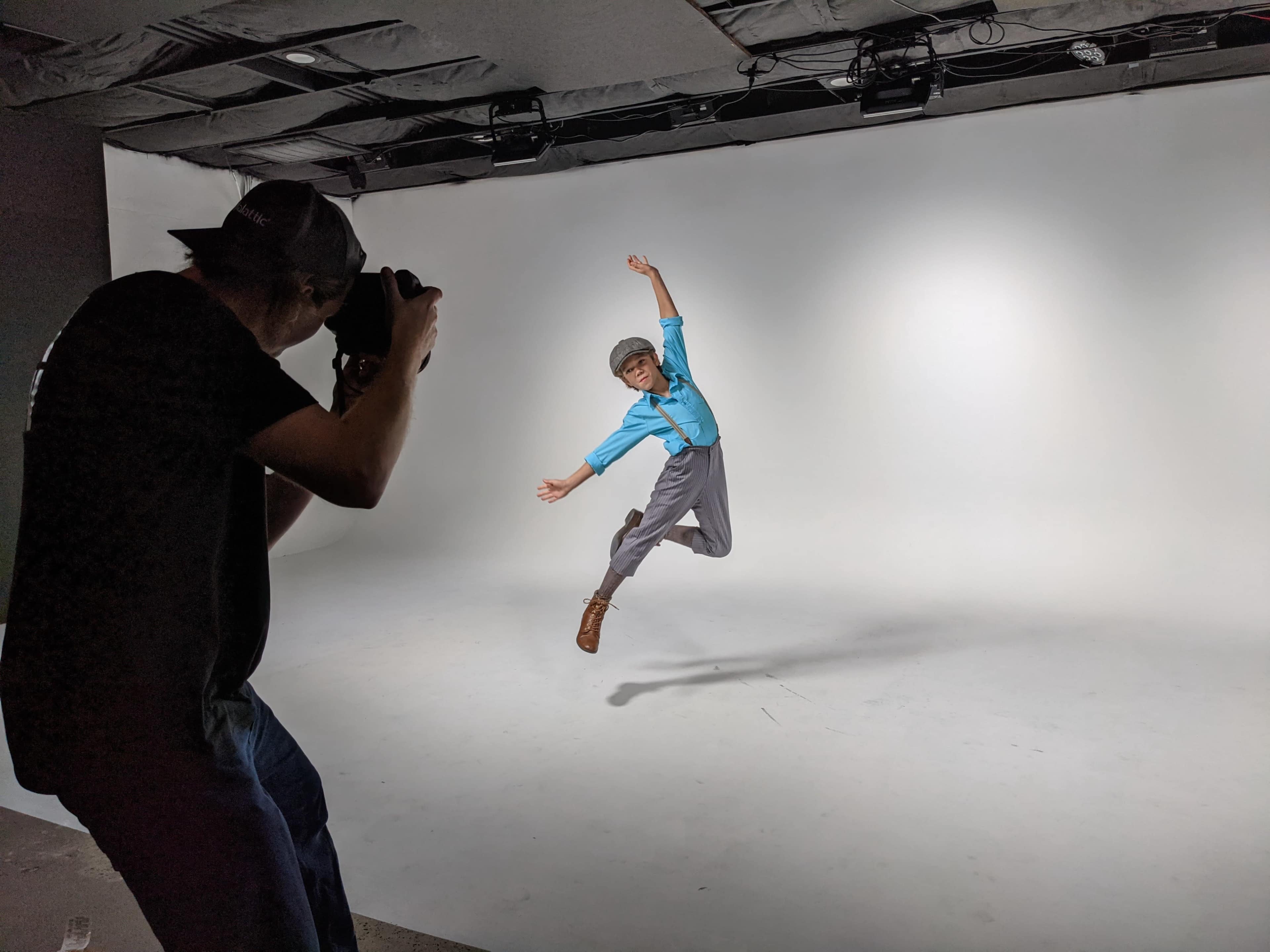 A child wearing a blue shirt and gray pants jumps in a photography studio while a photographer captures the moment.
