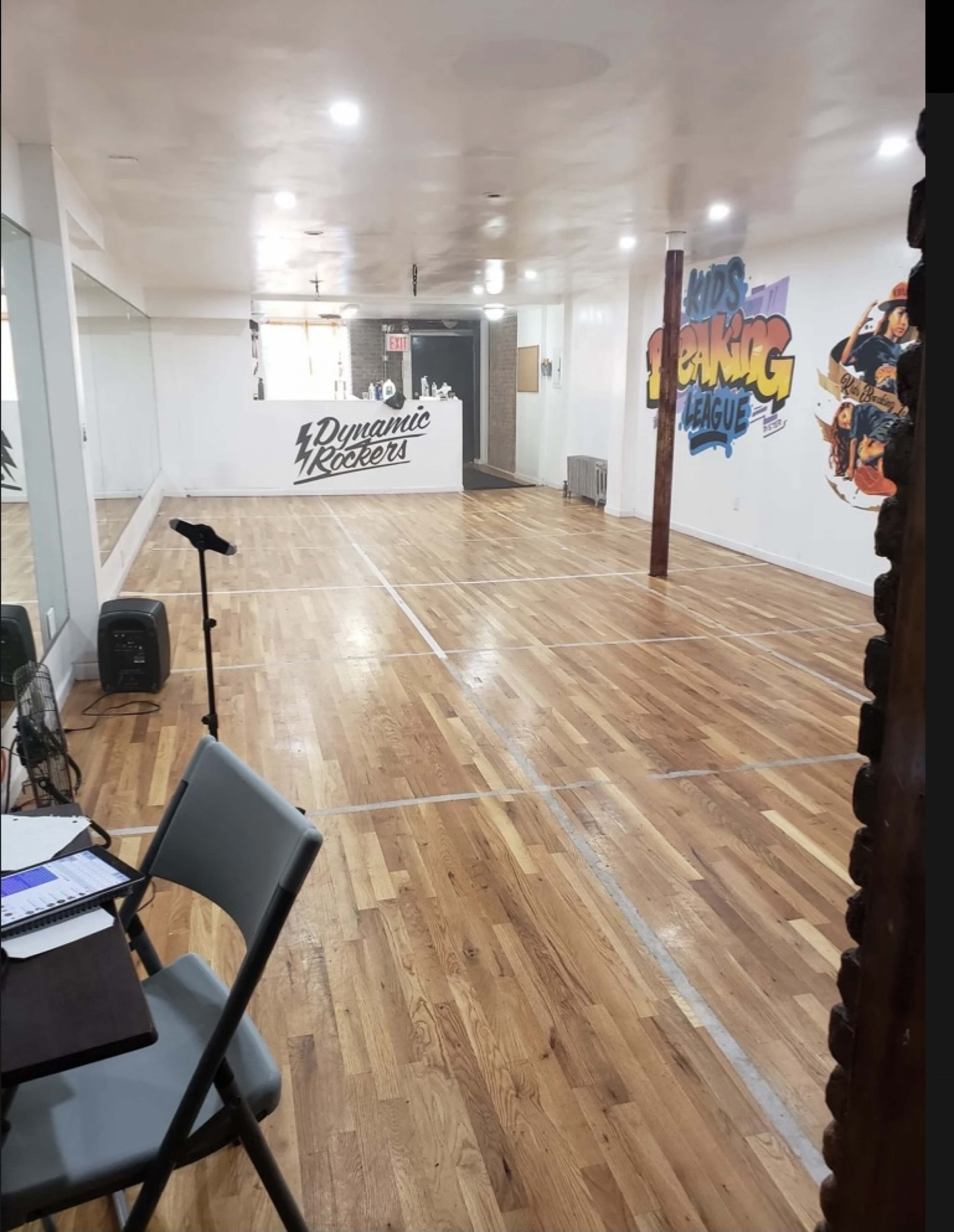 A spacious room with wooden flooring, featuring a microphone on a stand, a table with chairs, and a wall mural that reads "Kids Speaking League."