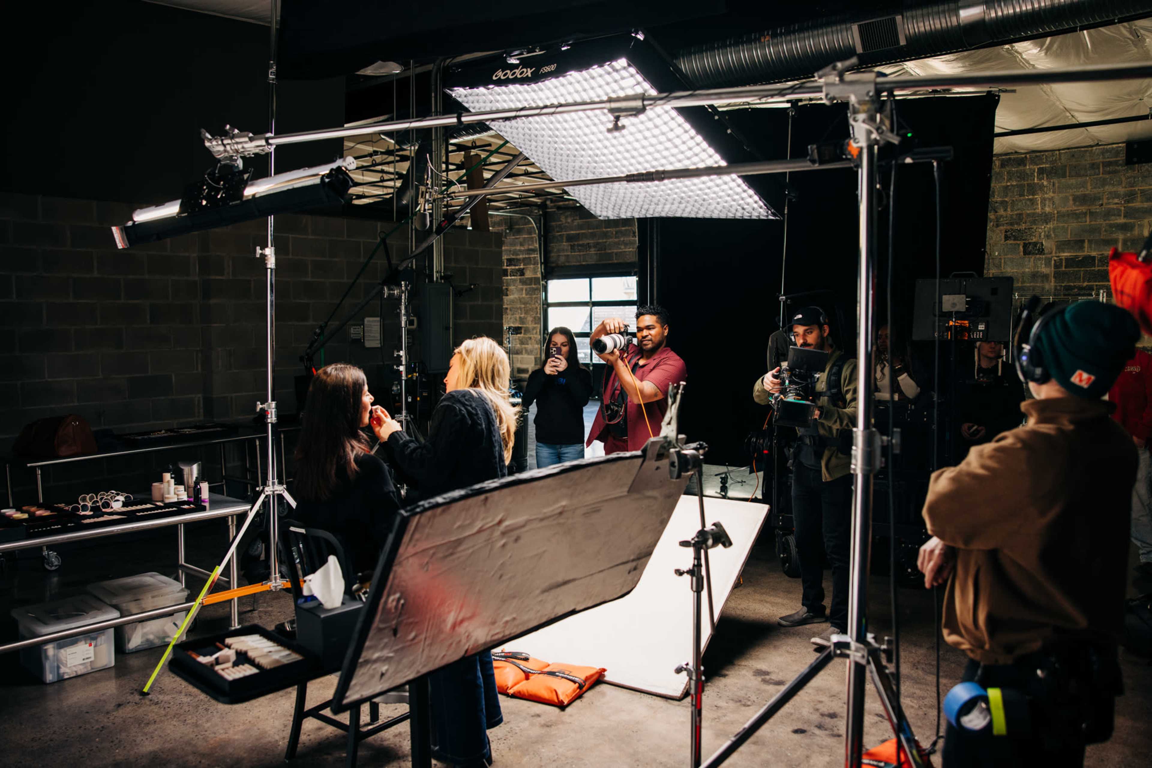 A group of people is engaged in a photo shoot in a studio, with a photographer capturing a model while a makeup artist applies touch-ups.