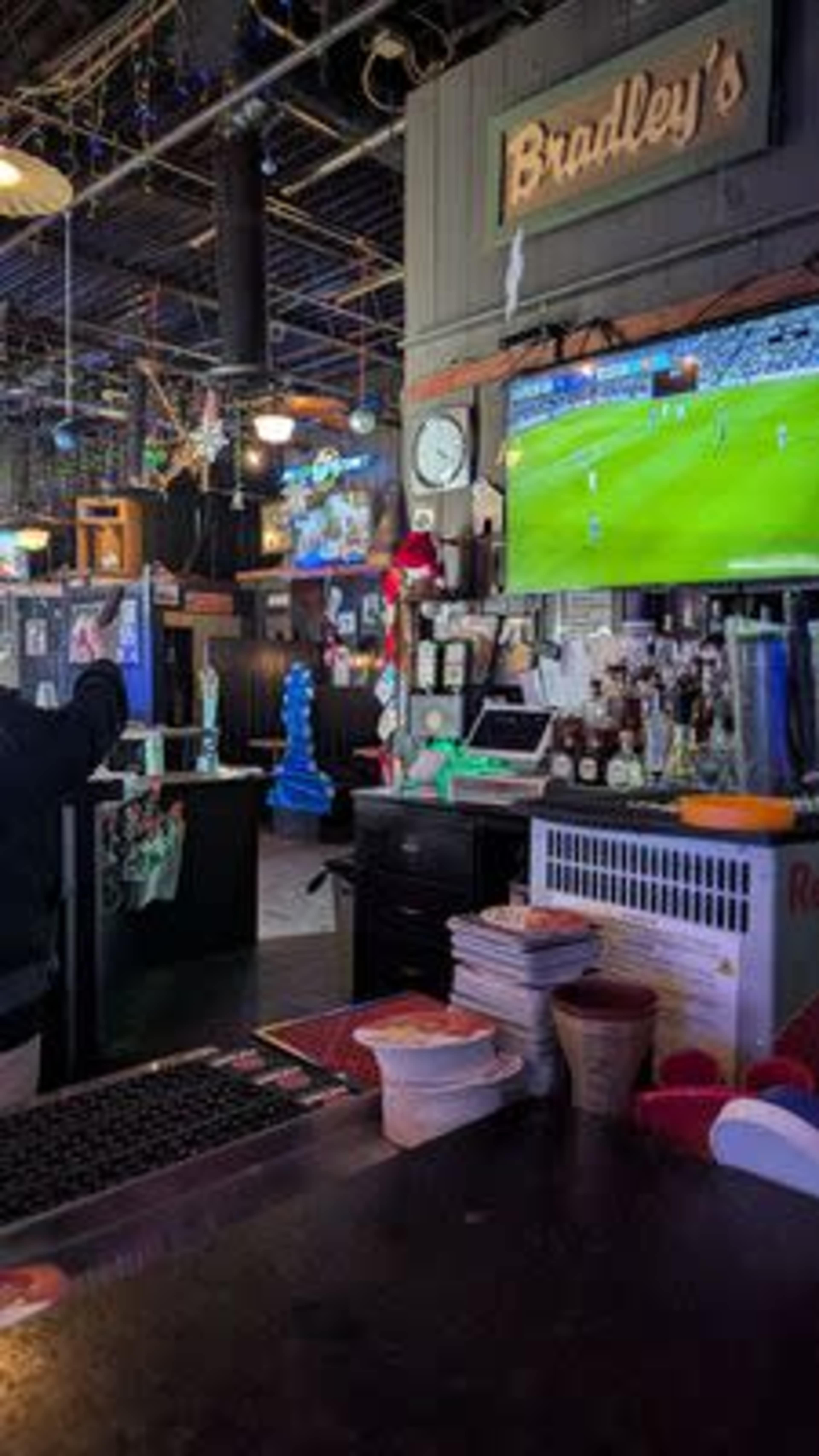 The interior of a bar named "Bradley's" features a dark wooden counter, various bottles on display, and multiple televisions showing a sports game.