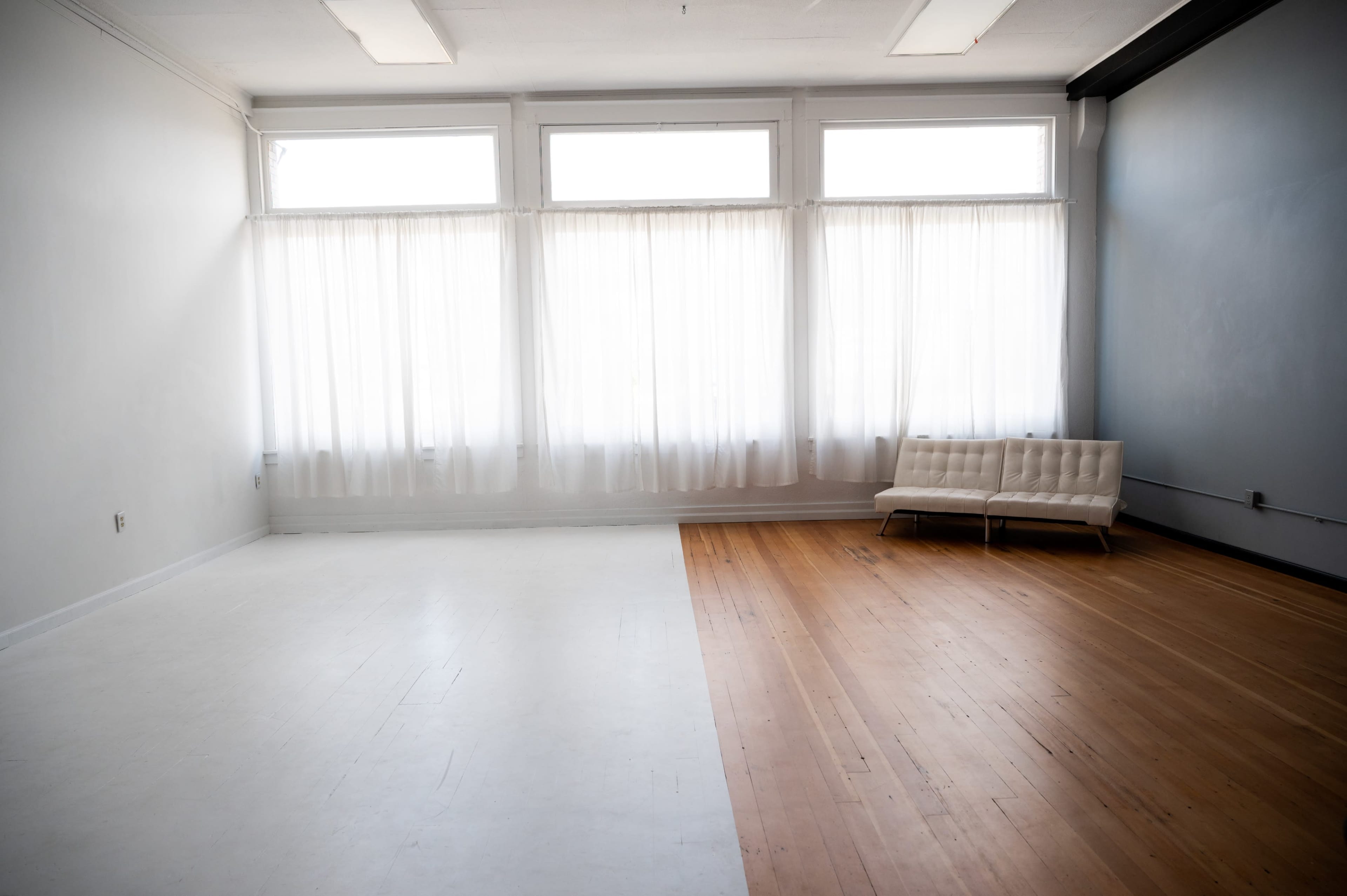 A bright, empty room featuring large windows with sheer curtains on one side and a couch placed on a wooden floor on the other side.