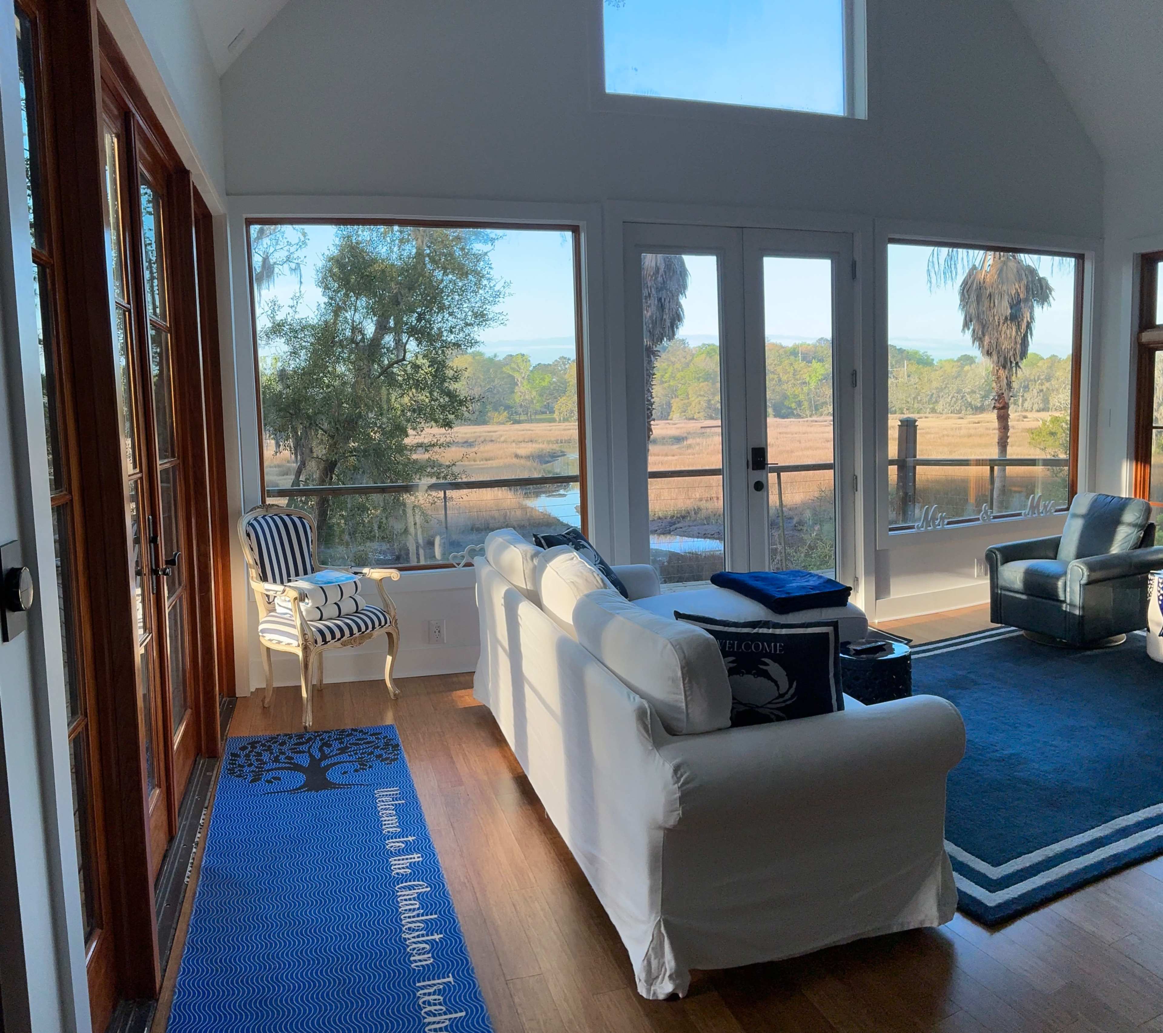 A bright living room with large windows overlooks a calm marsh area, featuring a white sofa, a blue carpet, and a striped chair.