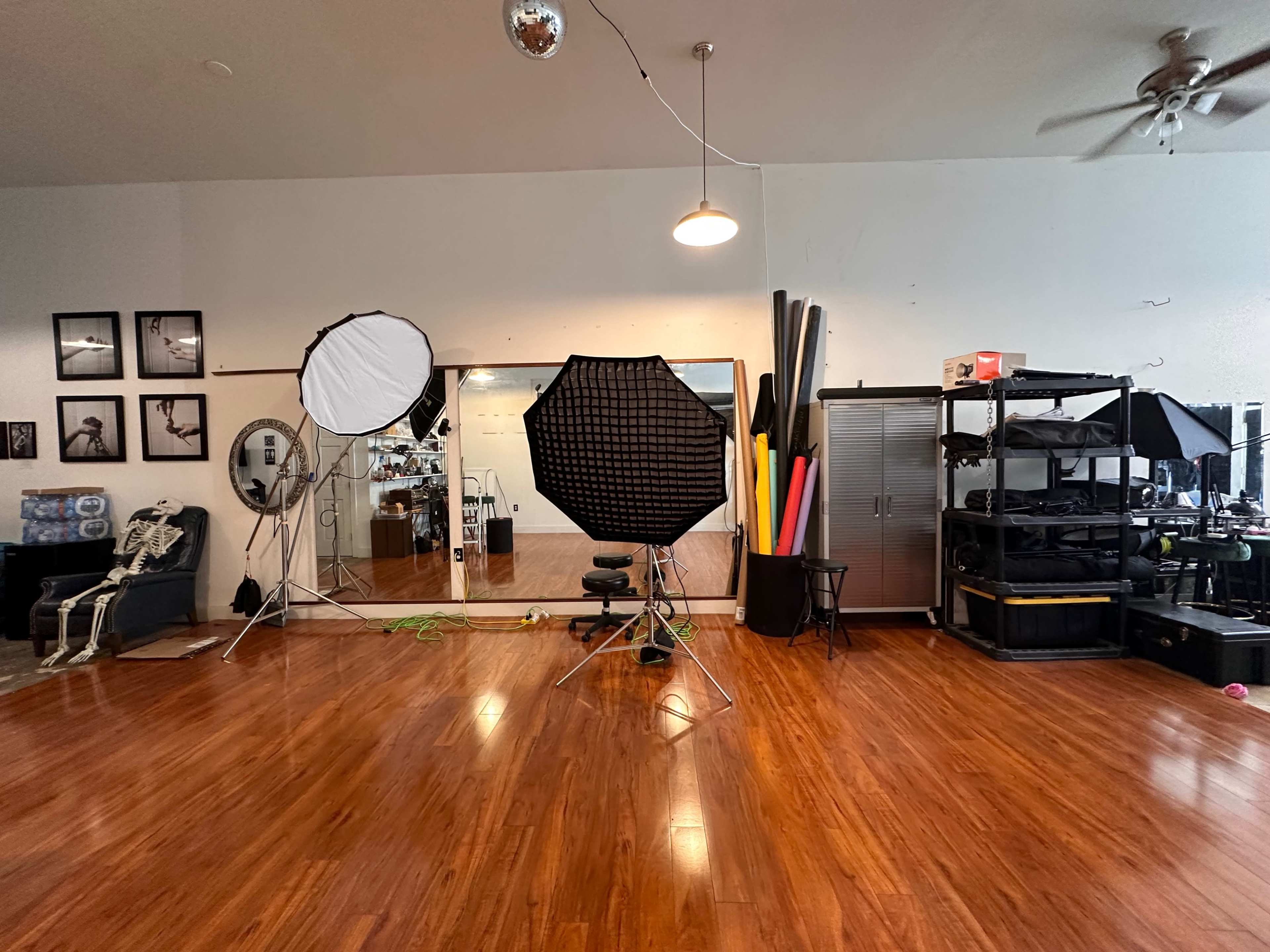 The image shows a photography studio with large lighting equipment, a mirror, and various props arranged in a spacious, wooden-floored room.
