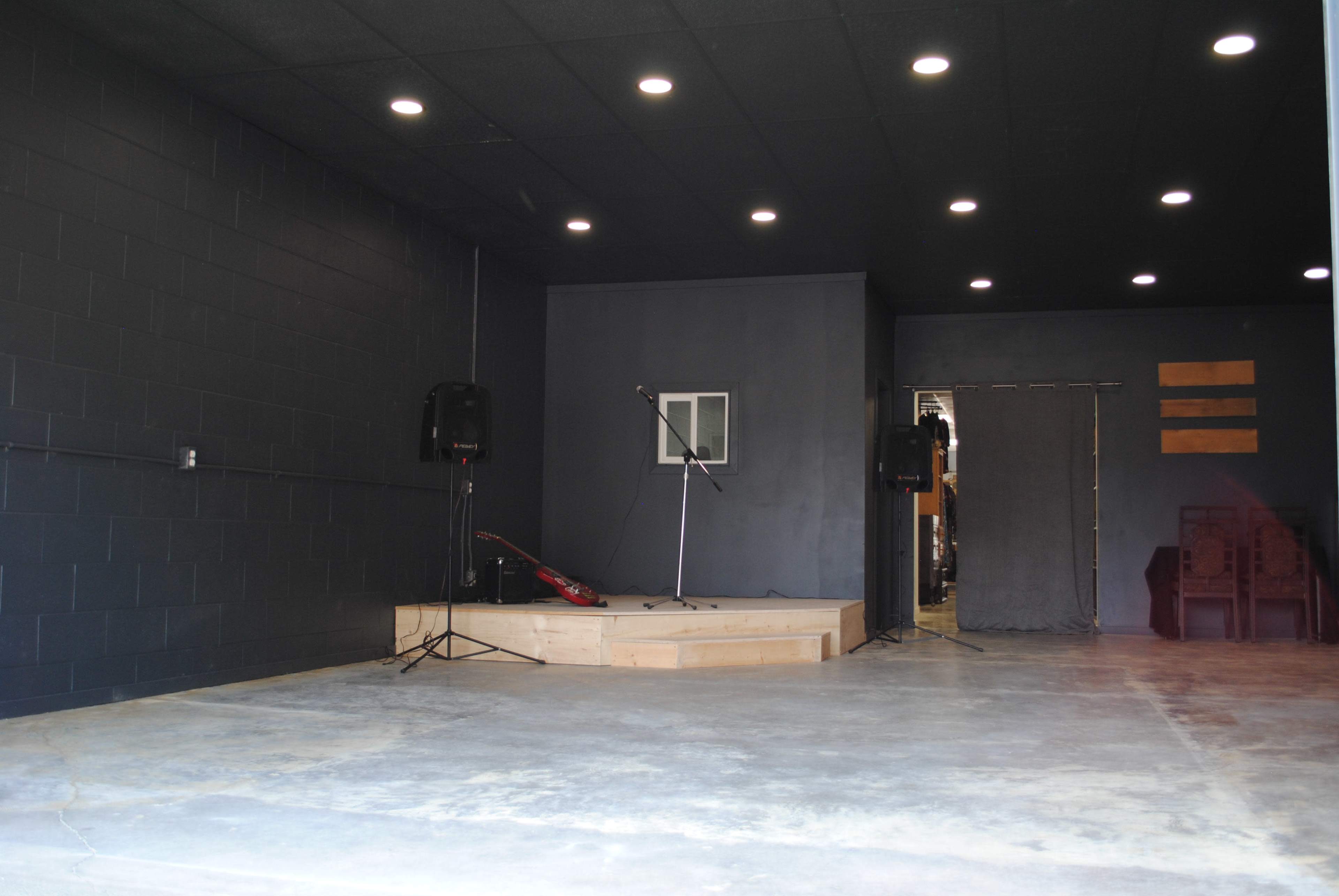 The image shows a dimly lit performance space with a raised wooden stage, a microphone stand, and speakers on either side, leading to an entrance in the background.