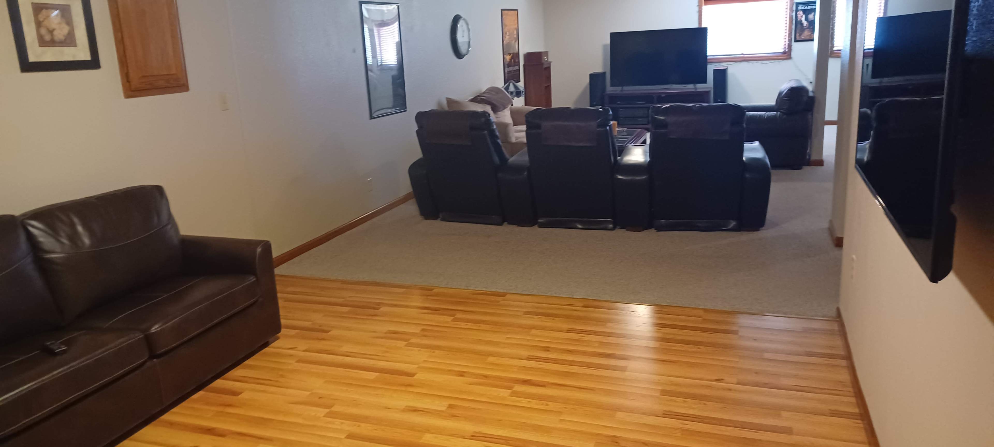 The image shows a living room with a wooden floor, a brown couch on the left, and a seating area with black recliners and a television in the background.