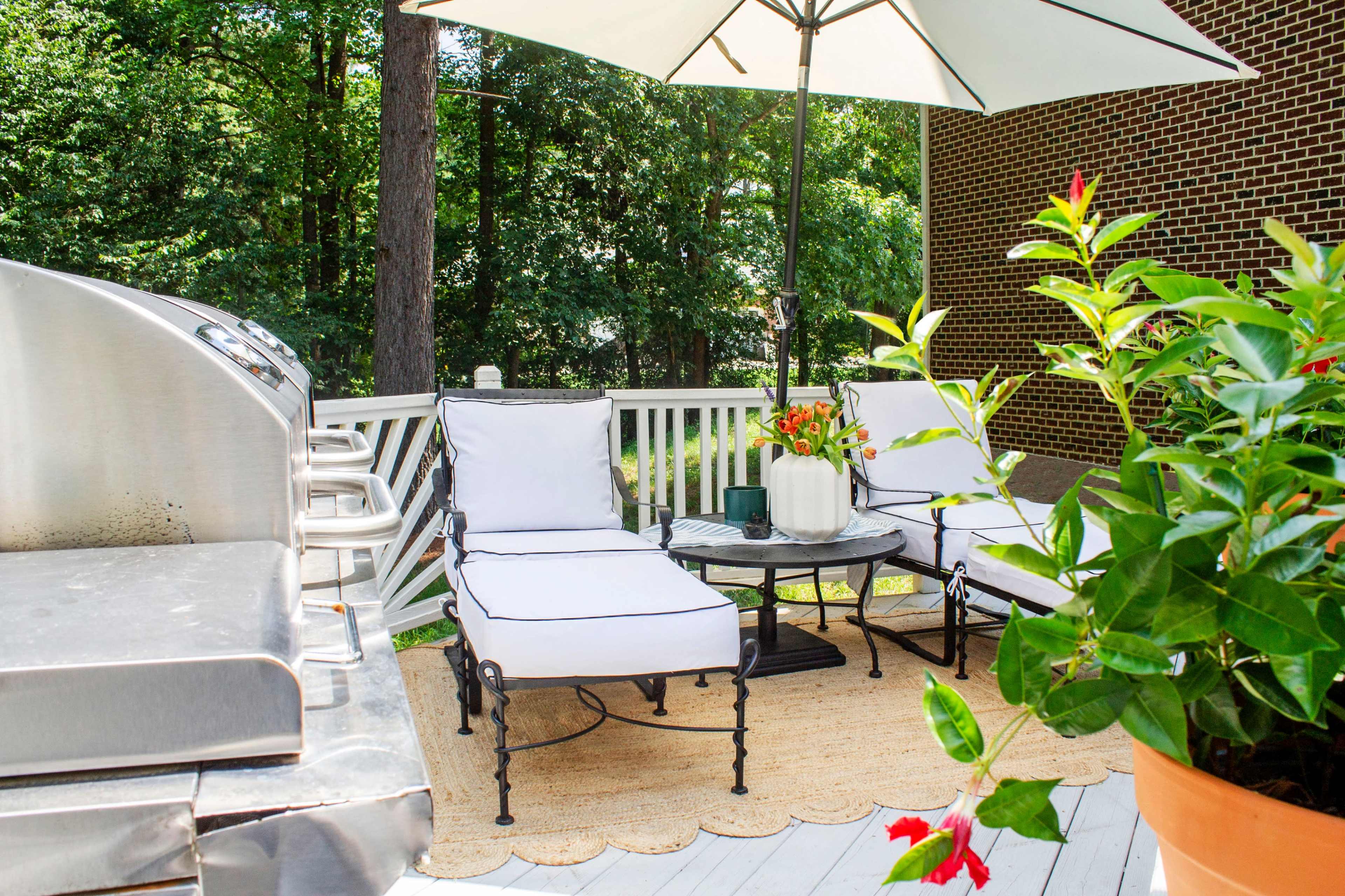 A patio area features two white lounge chairs, a round table, and a barbecue grill under an umbrella surrounded by greenery.