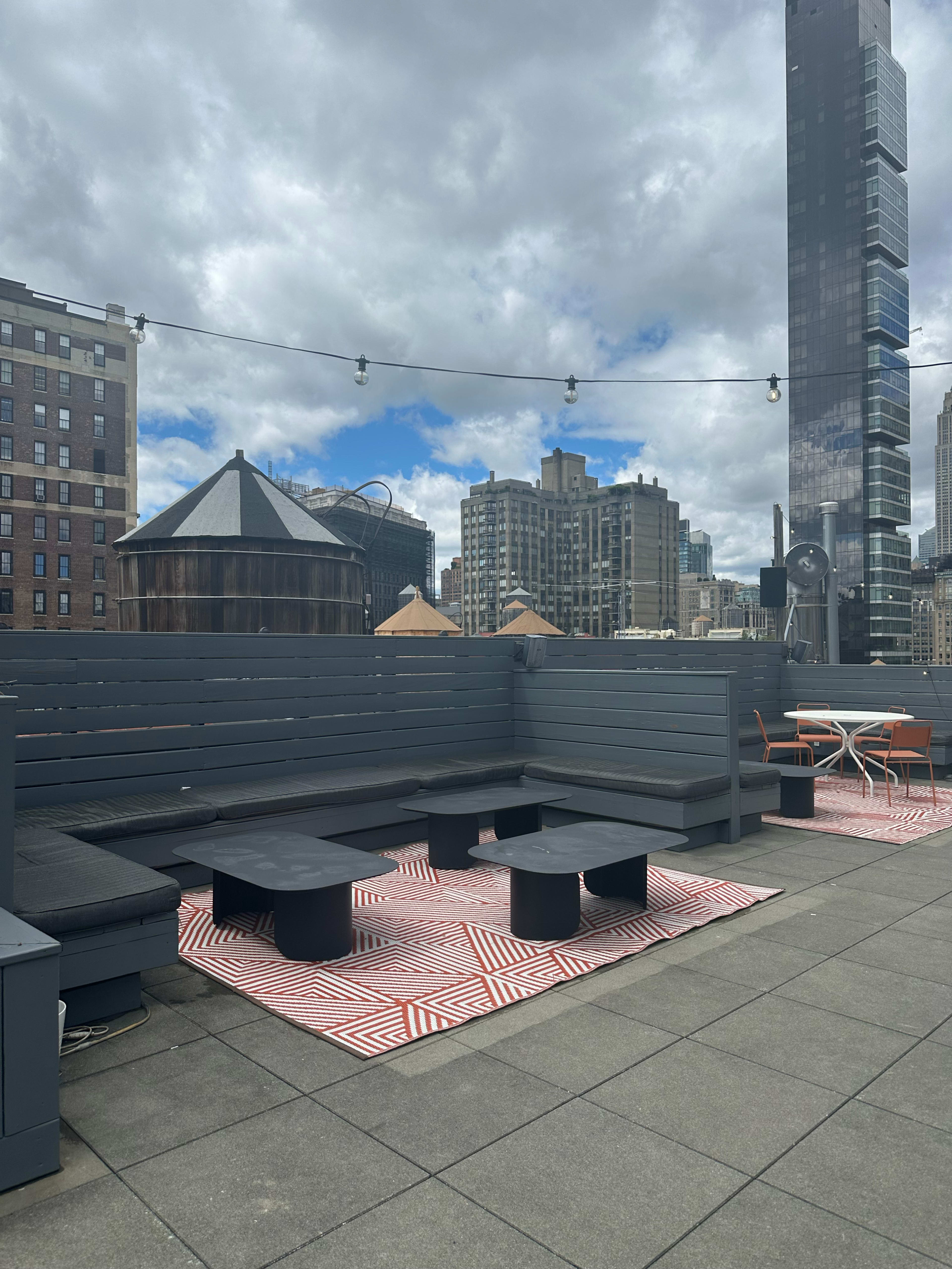 The image shows a rooftop lounge area with gray seating, black tables, and geometric-patterned rugs, overlooking a cityscape with tall buildings under a cloudy sky.