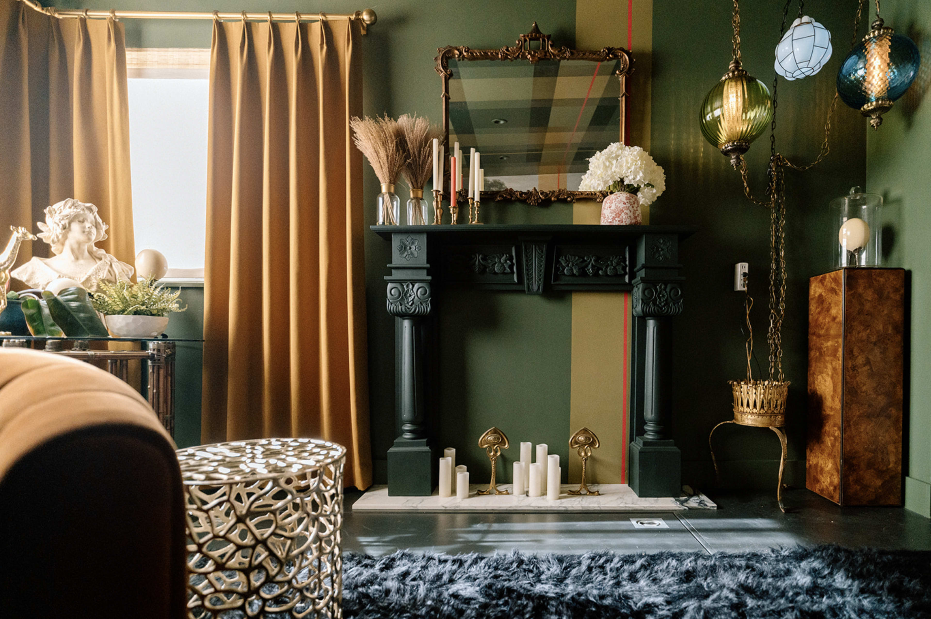 A stylishly decorated room featuring a dark green wall, an ornate fireplace with candles, a large mirror, amber curtains, and various decorative elements including plants and unique lighting fixtures.