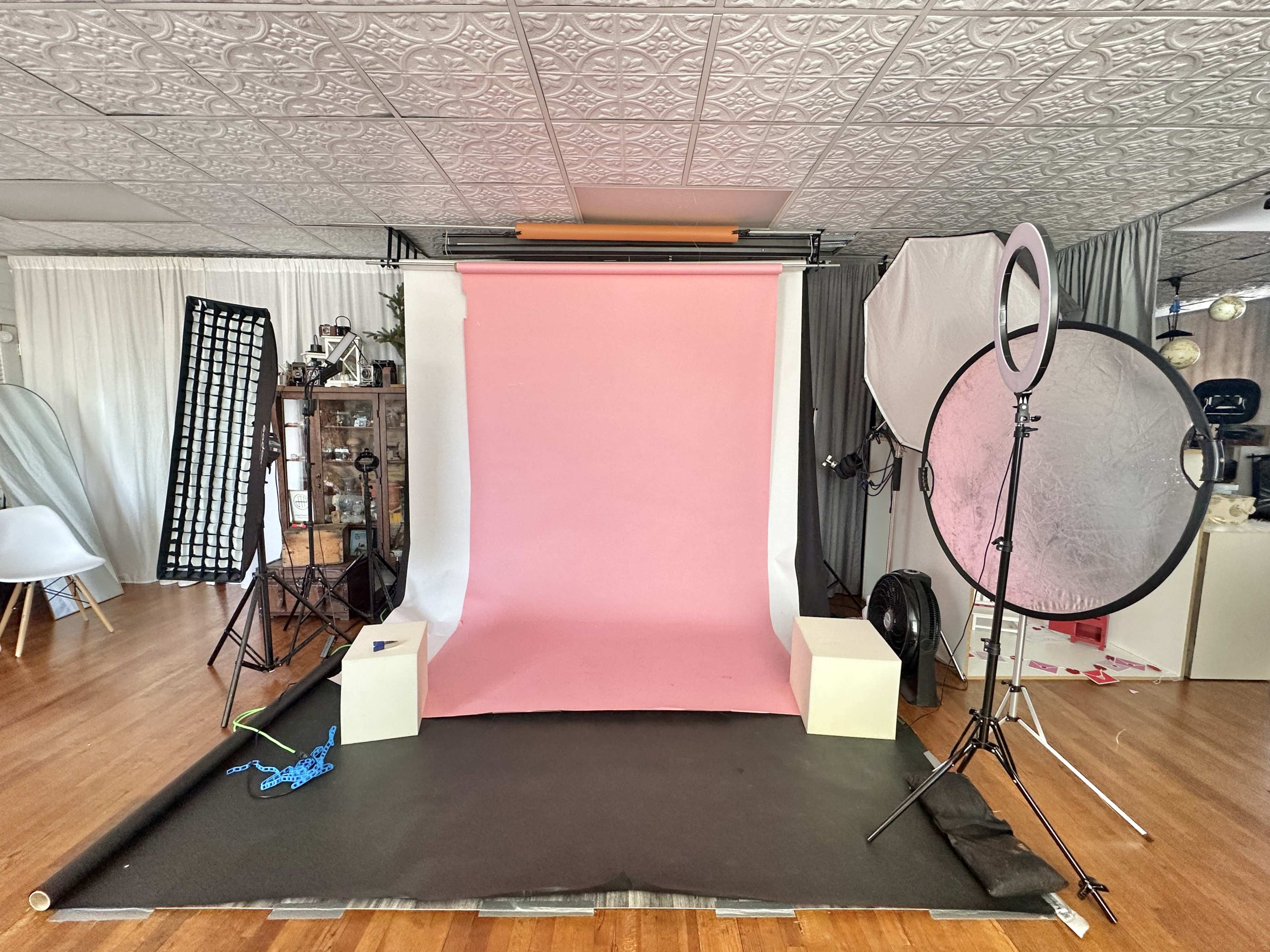 The image shows a photography studio set up with a pink backdrop, lighting equipment, and several props arranged in the space.