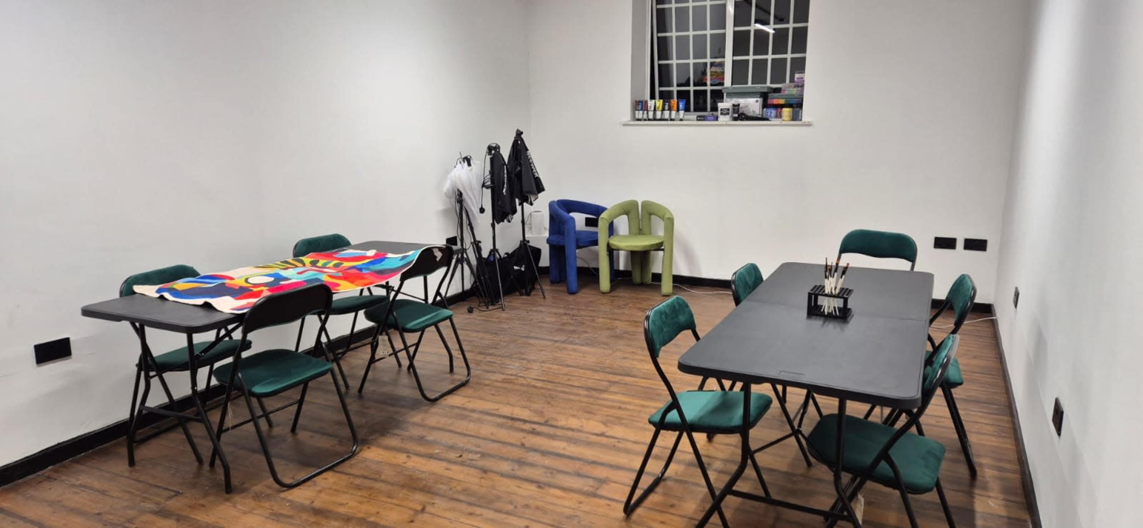 A spacious, empty room features two sets of folding tables surrounded by chairs, with a colorful cloth draped over one table and a small clothing rack against the wall.