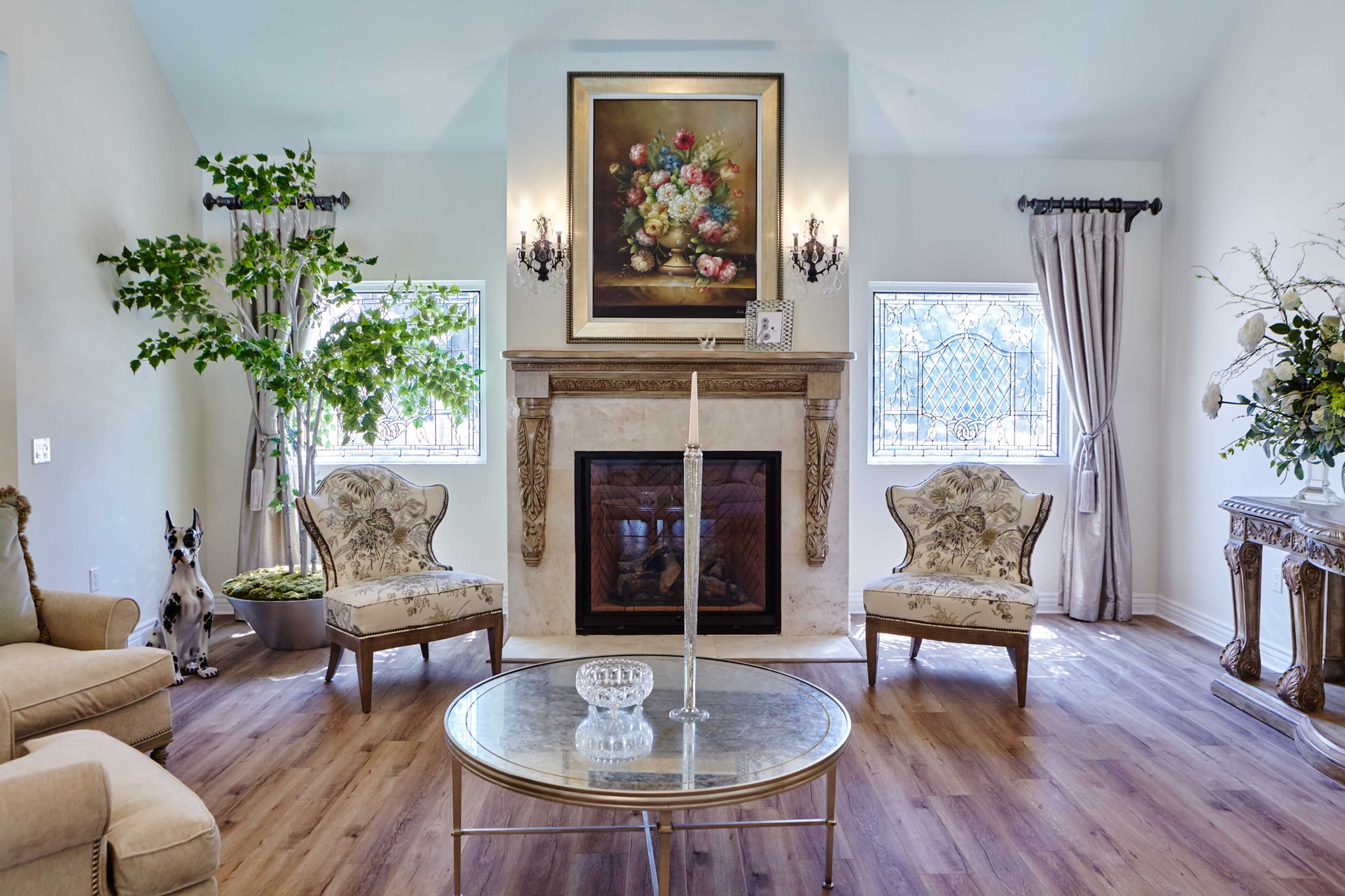 The living room features two ornate chairs, a coffee table, and a fireplace adorned with a large floral painting, surrounded by windows that allow natural light to enter the space.