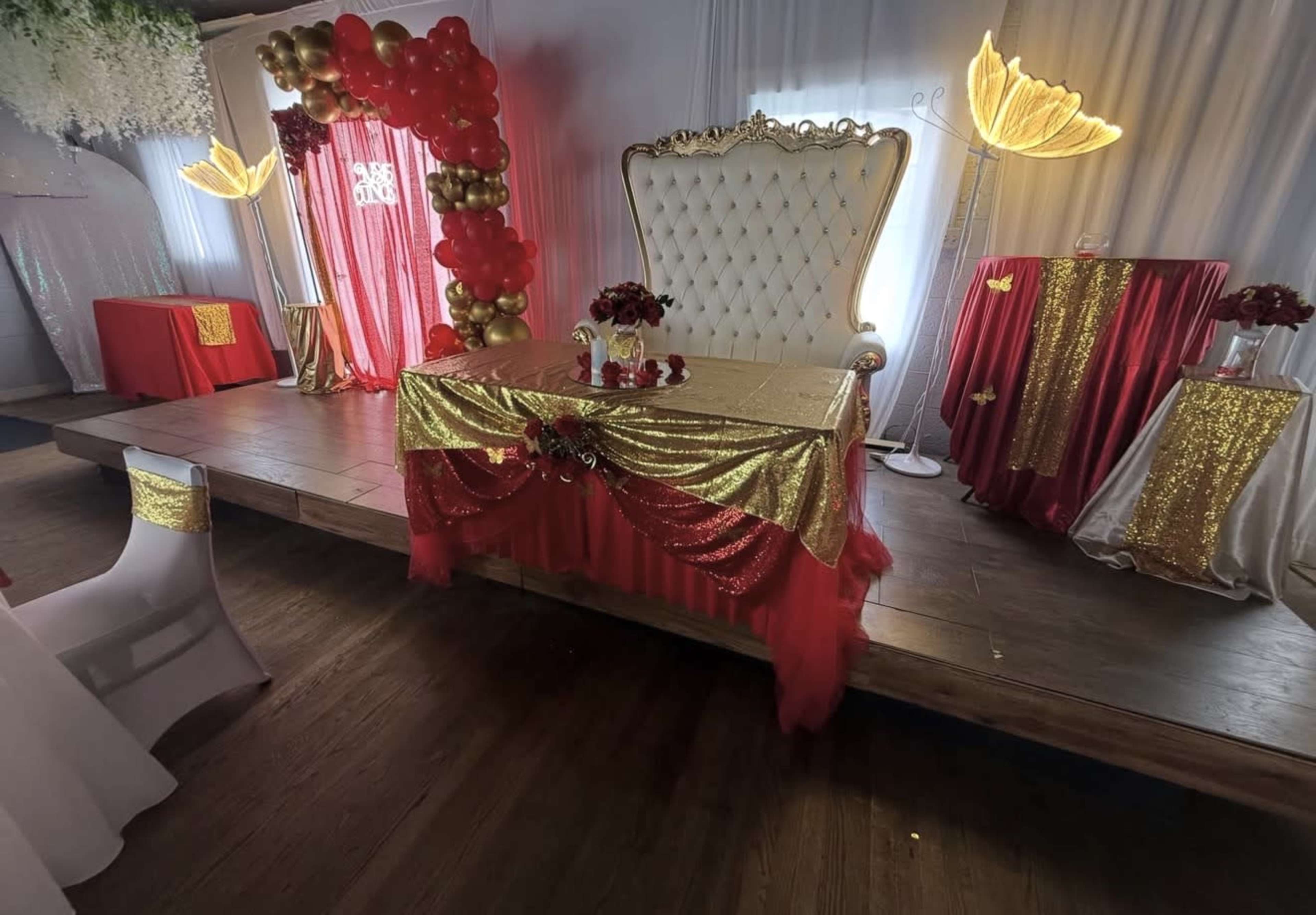 The image shows a decorated stage with a large white throne, a festive table covered in red and gold fabric, and various decorative elements including balloons and illuminated floral lamps.