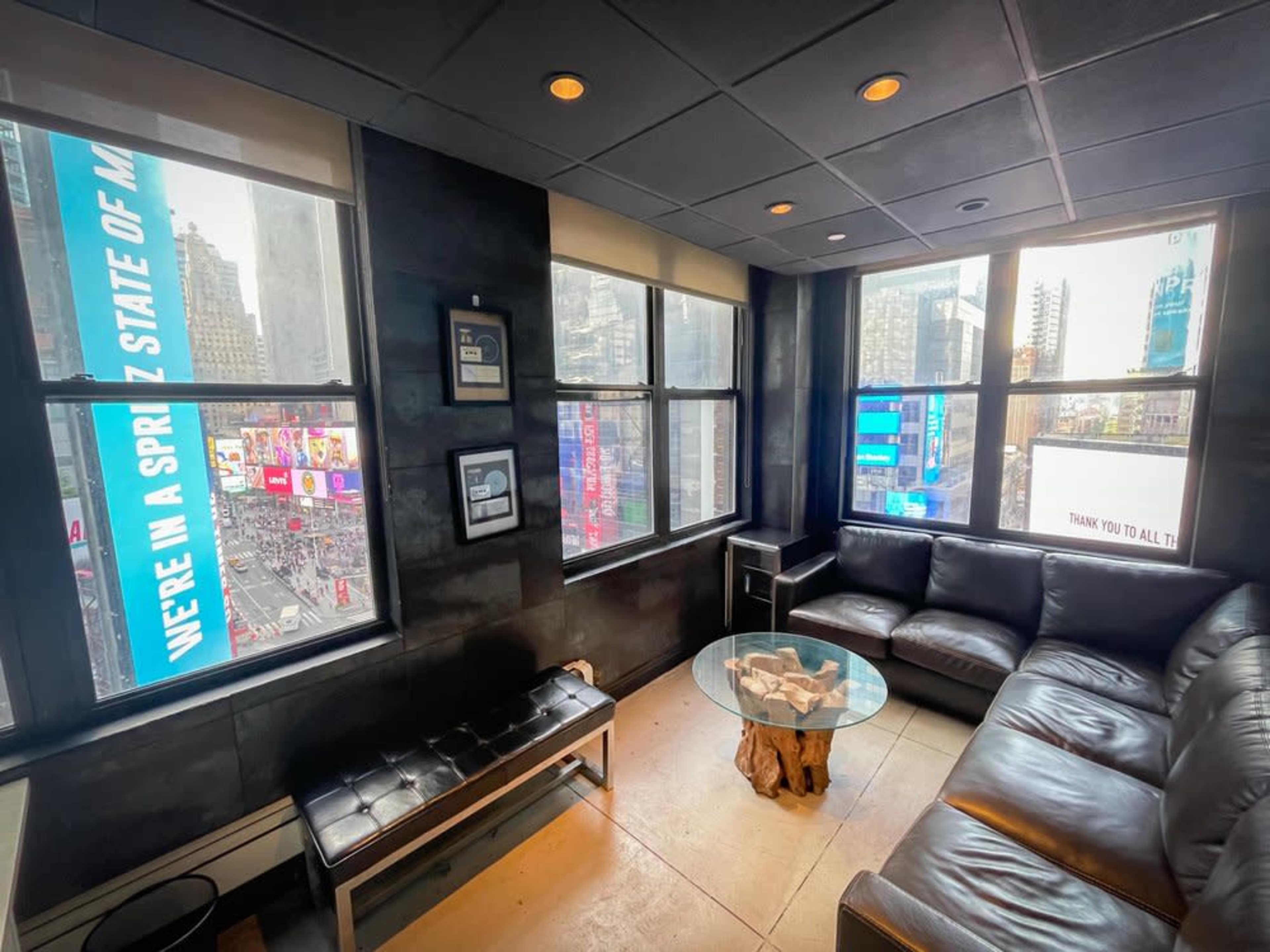 A modern lounge area features a black leather couch and a glass coffee table, with large windows showcasing the vibrant advertisements of Times Square.