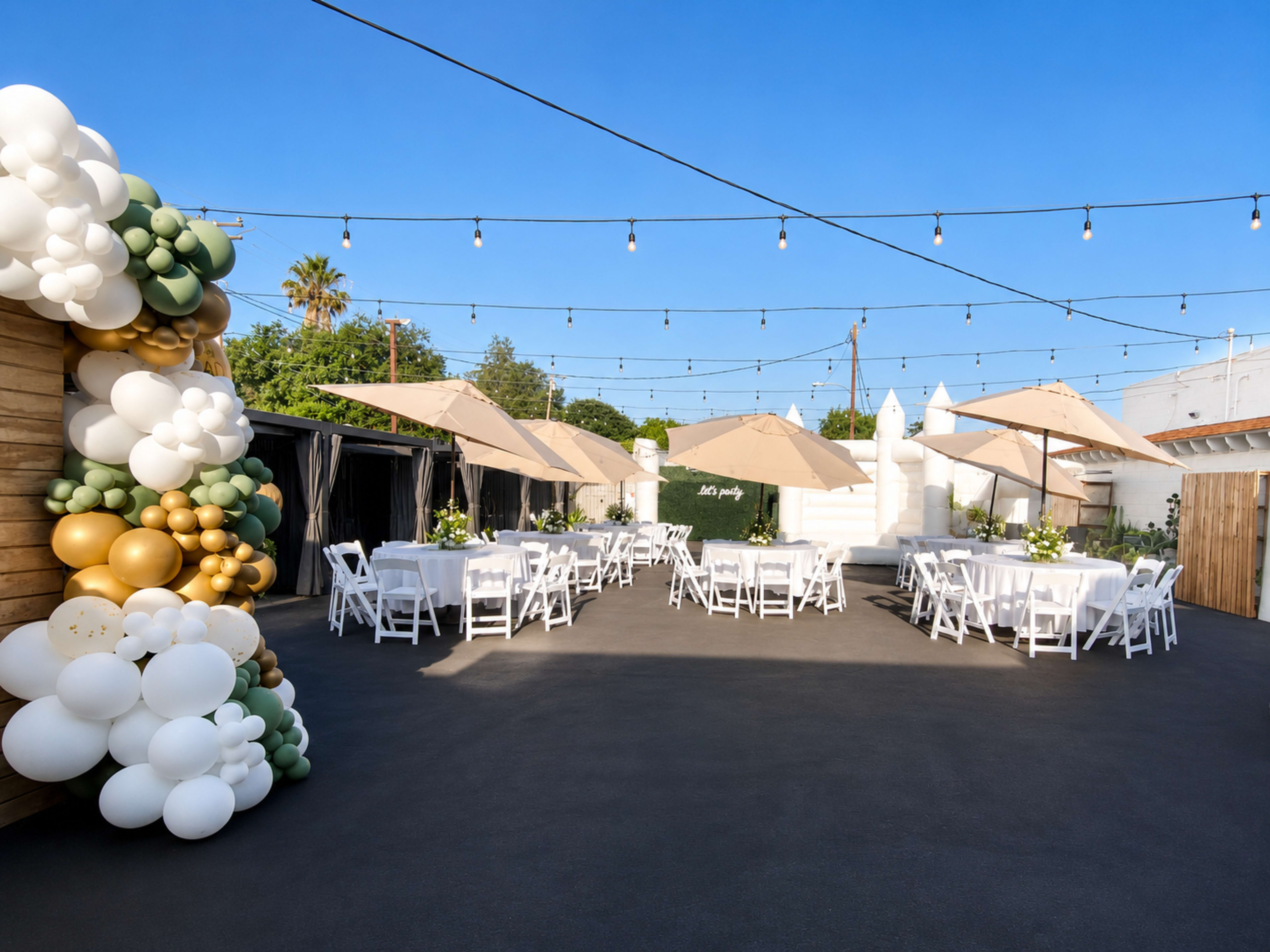 The image shows an outdoor event space featuring several round tables with white tablecloths, adorned with centerpieces, and large umbrellas for shade, surrounded by decorative balloons.