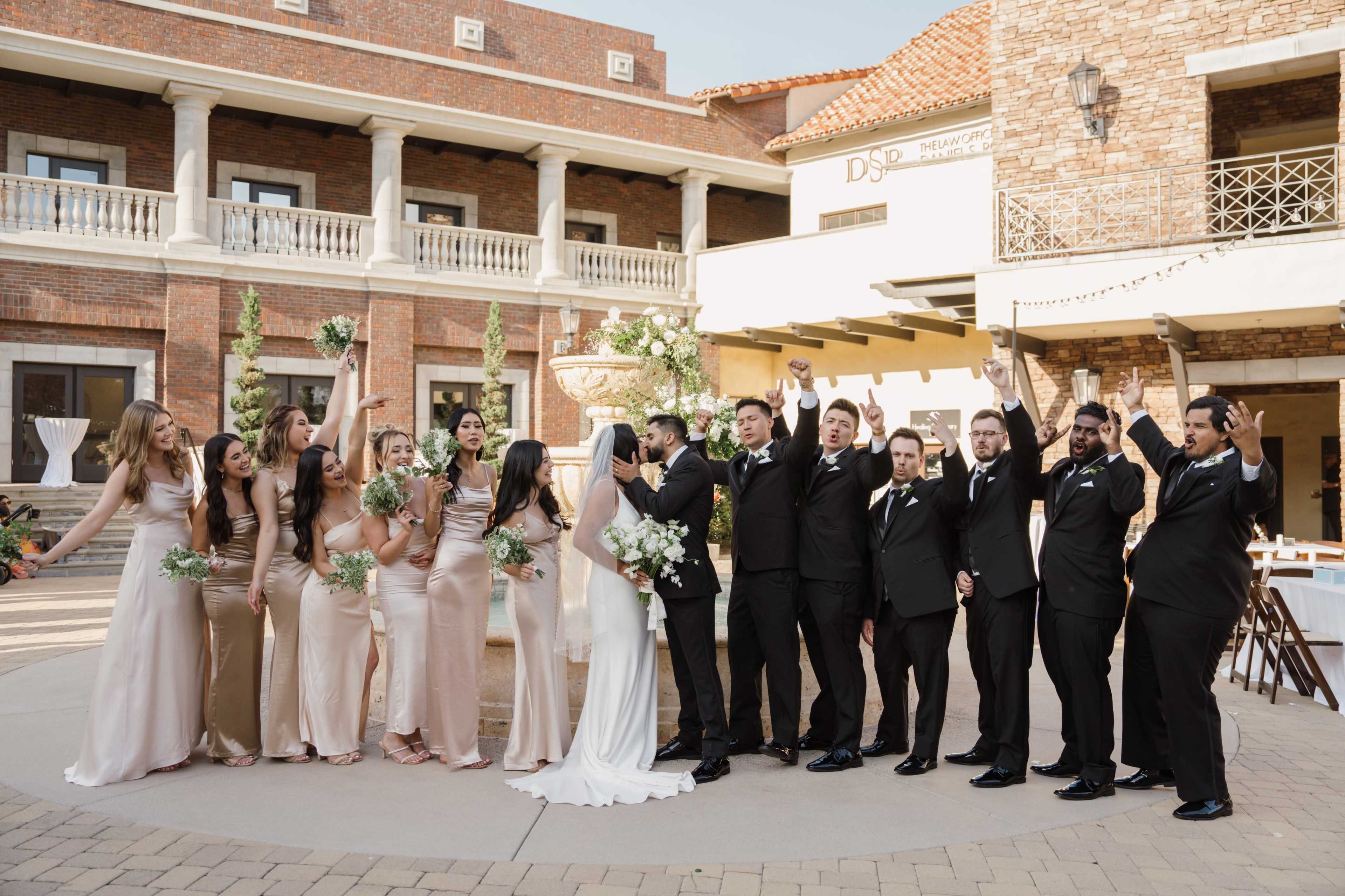 A group of wedding party members in formal attire pose together outdoors, celebrating near a fountain in a courtyard.